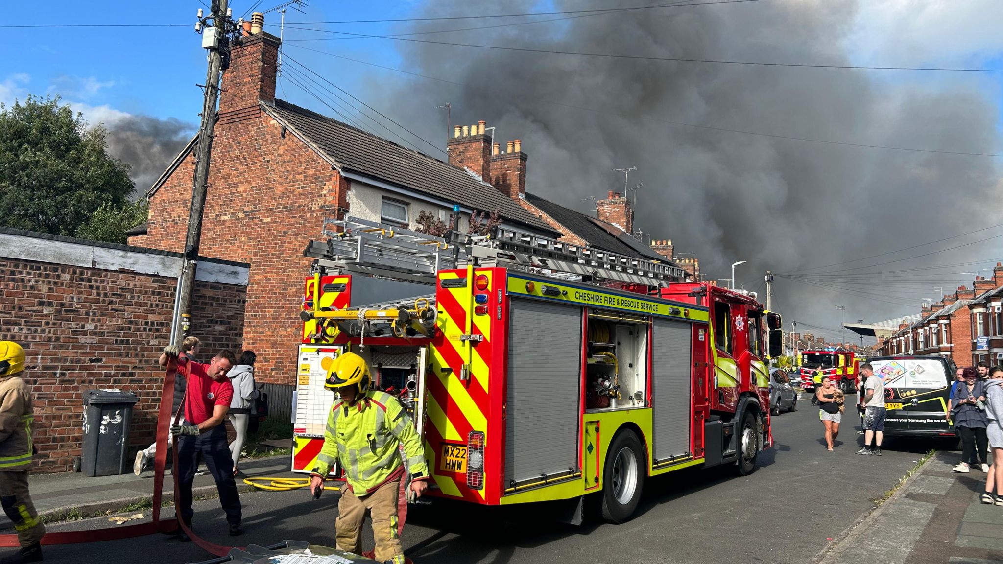 Firefighters set up a hose from a fire engine in the middle of a residential street as smoke billows behind buildings in the  background. 