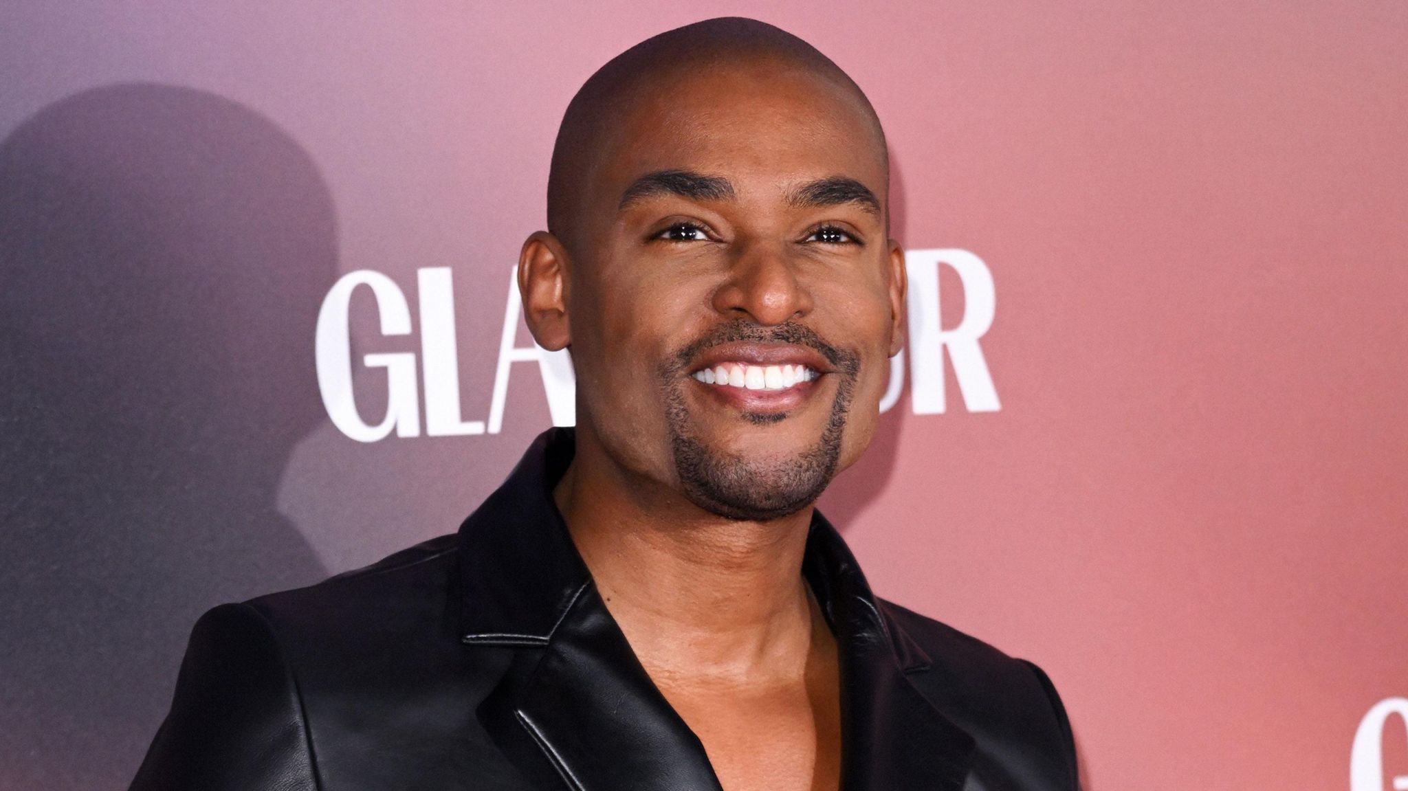 Paul Brunson pictured on a red carpet. Paul is a black man in his 40s with a shaved heard and short goatee style beard. He wears a black blazer with no shirt and smiles at the camera. 