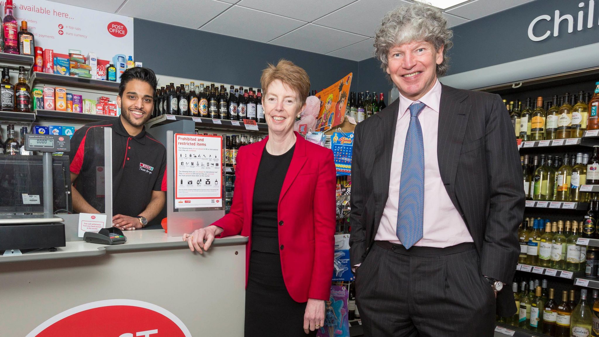 Than Thevarajah stands behind the till in his post office to the right of smiling Paula Vennells and Tim Parker with shelves stocked with alcohol in the background