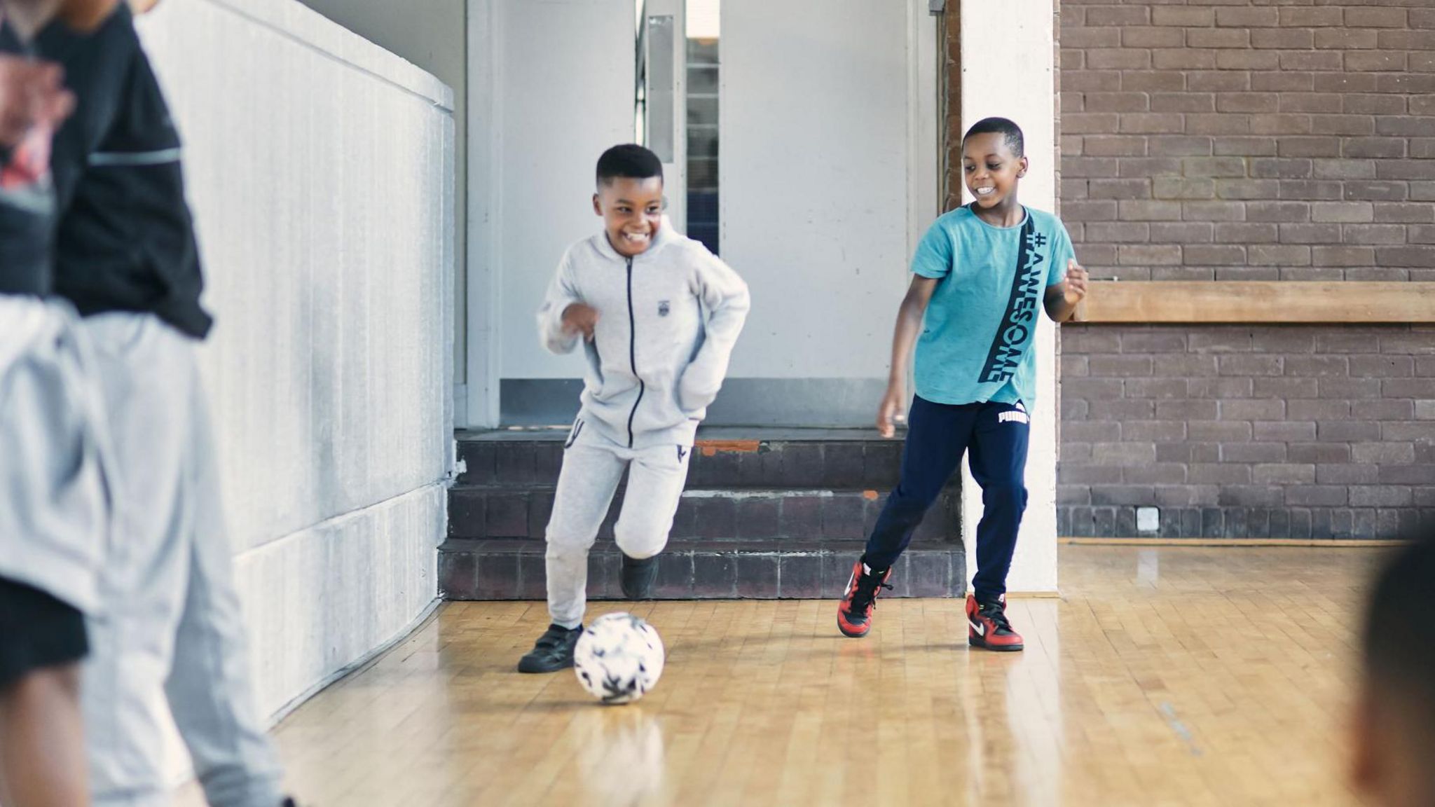 Two boys playing football inside, one wearing a pale tracksuit and smiling as he's running with the ball in front and another child wearing a blue tshirt with the words awesome on it is alongside, smiling. 