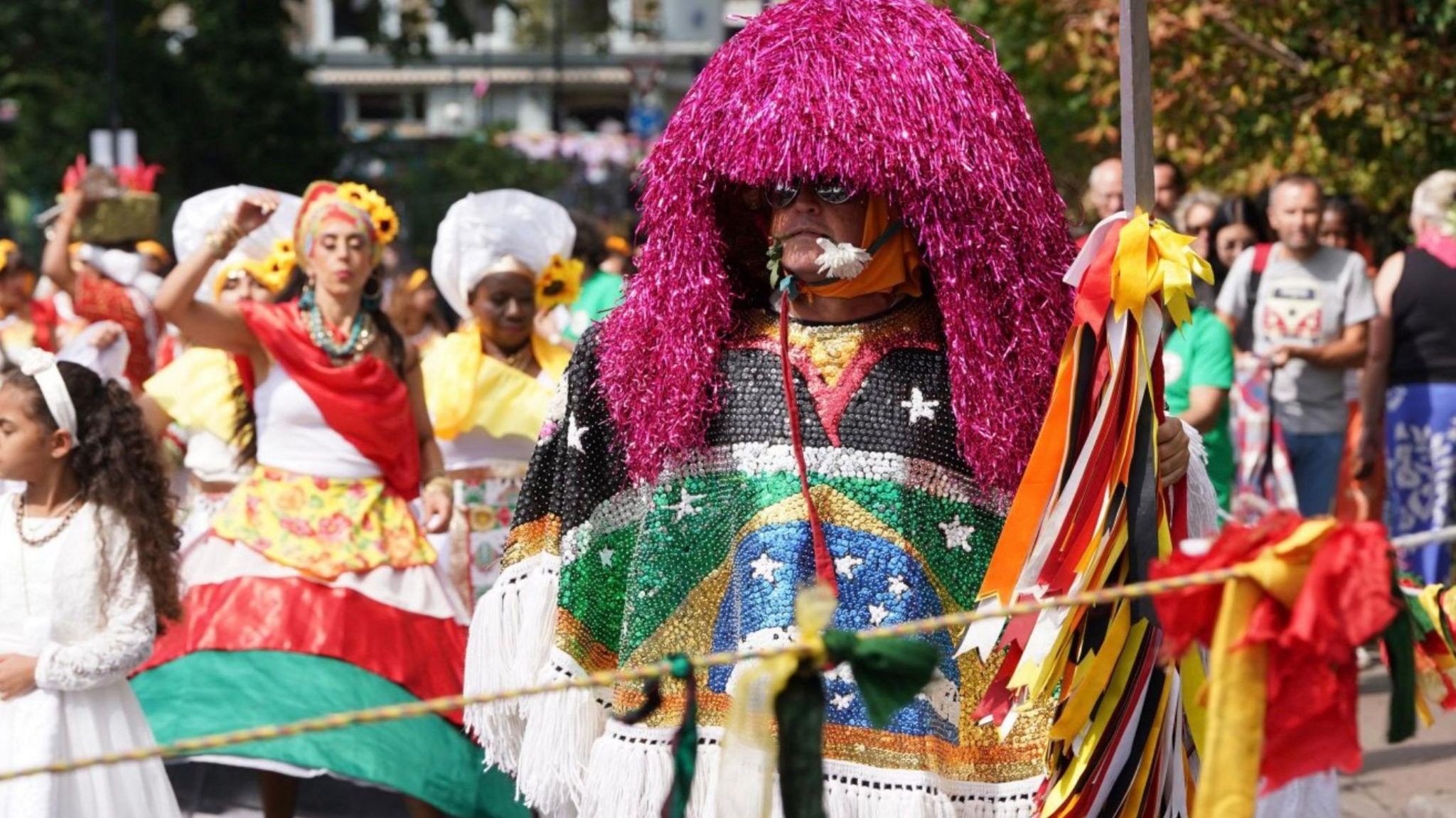 A man dresses up with colourful sequins which make up a Brazilian flag. He's also wearing a purple-streamed headdress and has a white flower in his mouth.