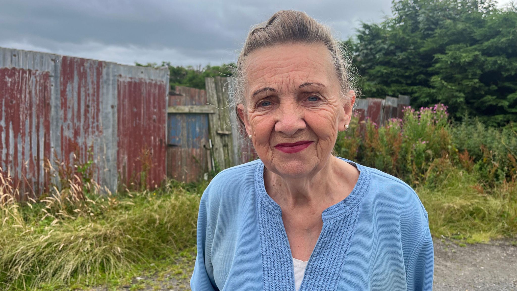 A woman with grey hair, wearing a light blue top and standing in front of a corrugated iron fence.