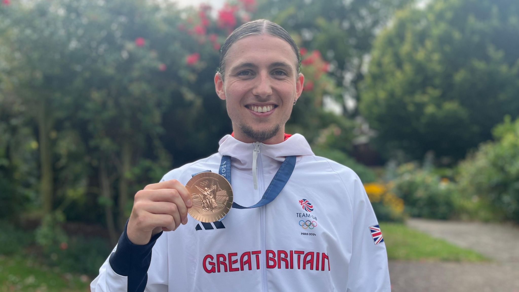 Lewis Davey wearing his white Great Britain tracksuit. His bronze medal hangs around his neck and he is holding it up with his right hand. The tracksuit has Great Britain in red writing across the middle. He is smiling proudly for the camera and has dark hair with a parting down the middle.