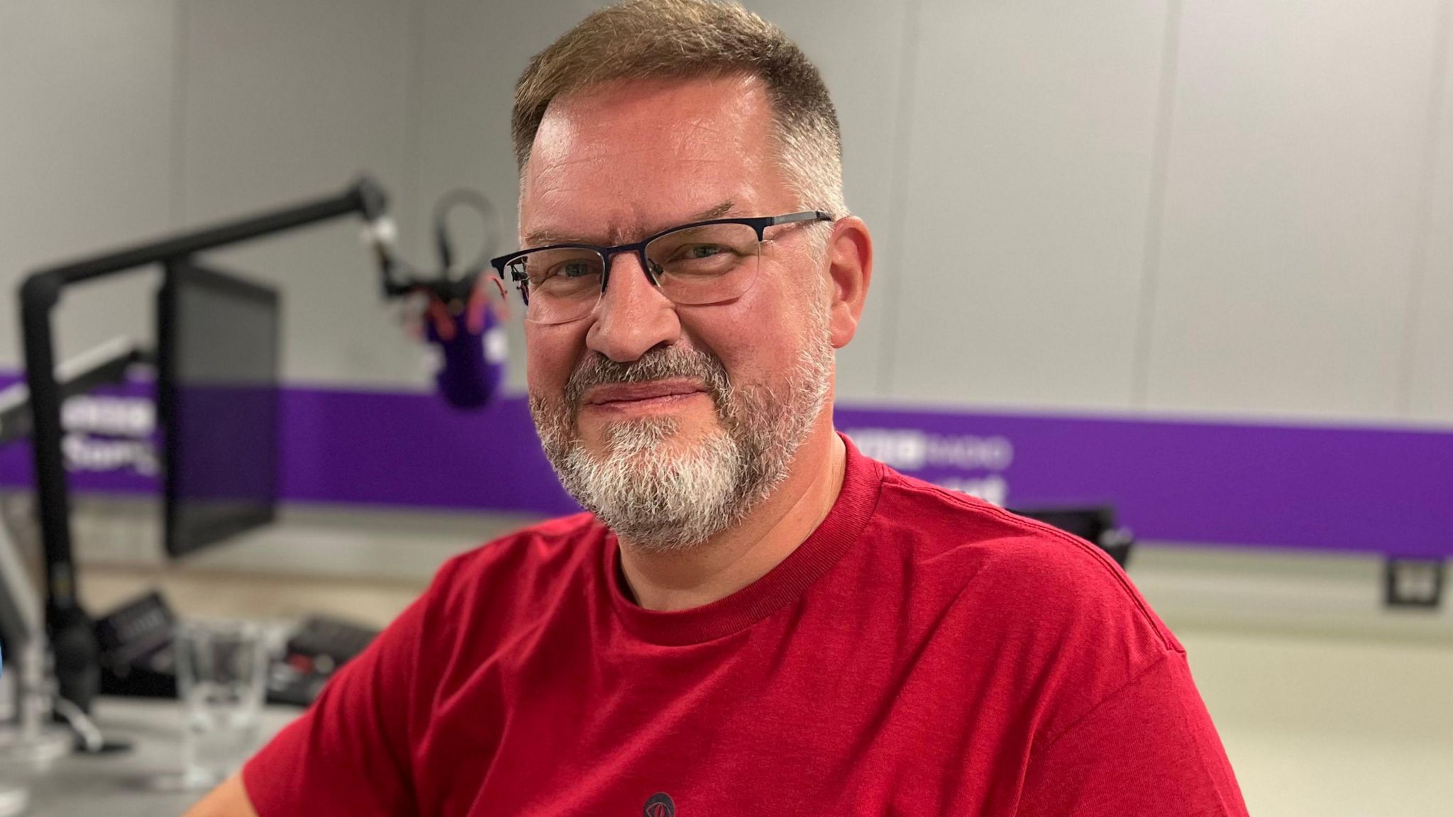 Jonathan Green. He is pictured in a radio studio, sat at a desk with a mic. Jonathan has short light hair and facial hair. He is wearing glasses and a red t-shirt. He is looking at the camera and smiling.