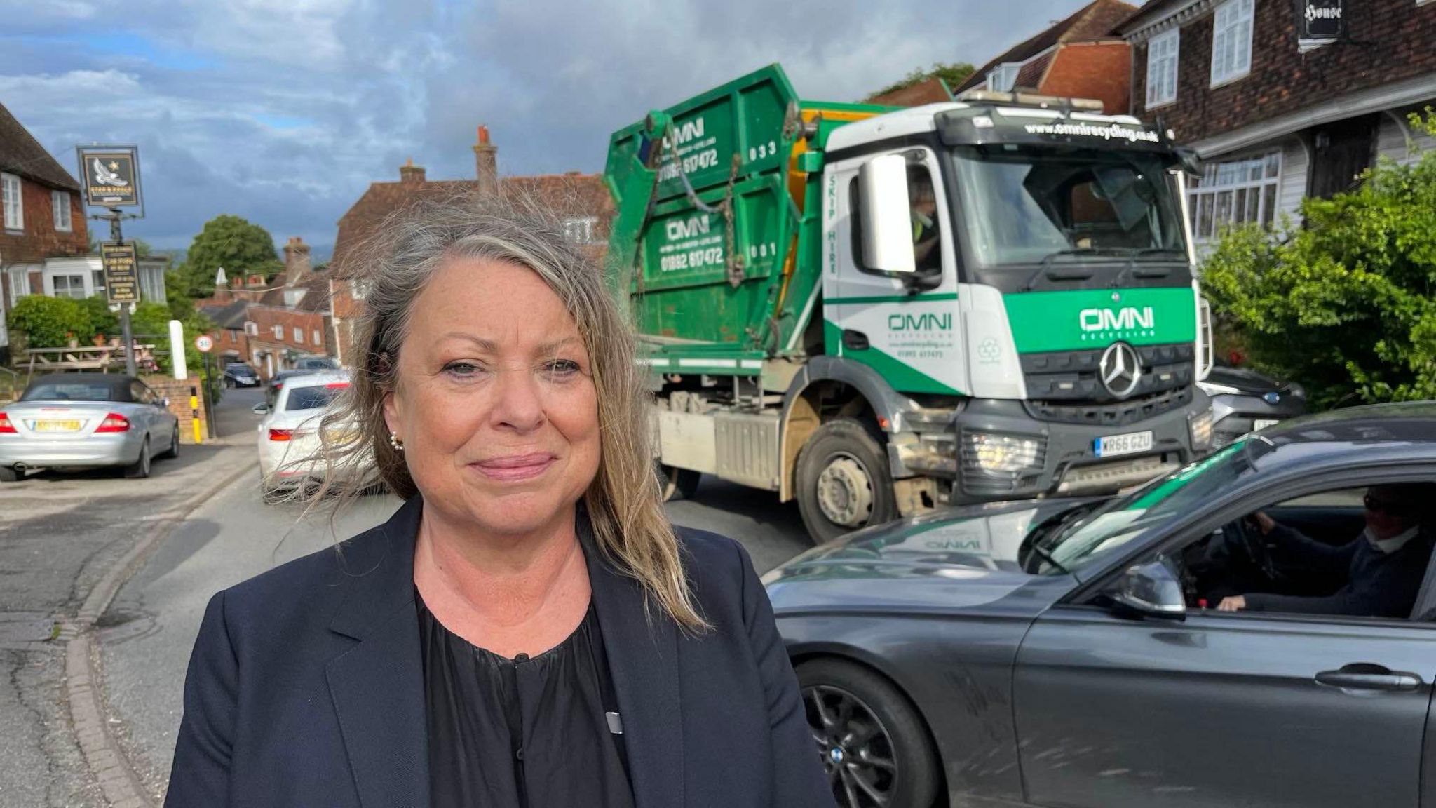 Grey haired woman stands at busy section of A262 with traffic squeezing past in background