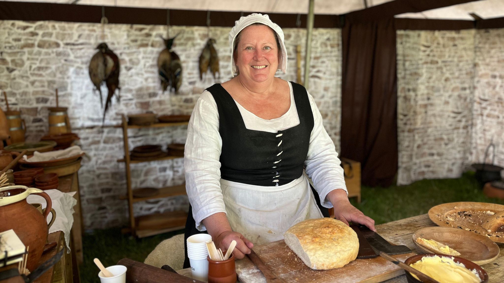 Tracey Grand looks into the camera dressed as Tudor cook with a fresh loaf of bread surrounded by historical cookware
