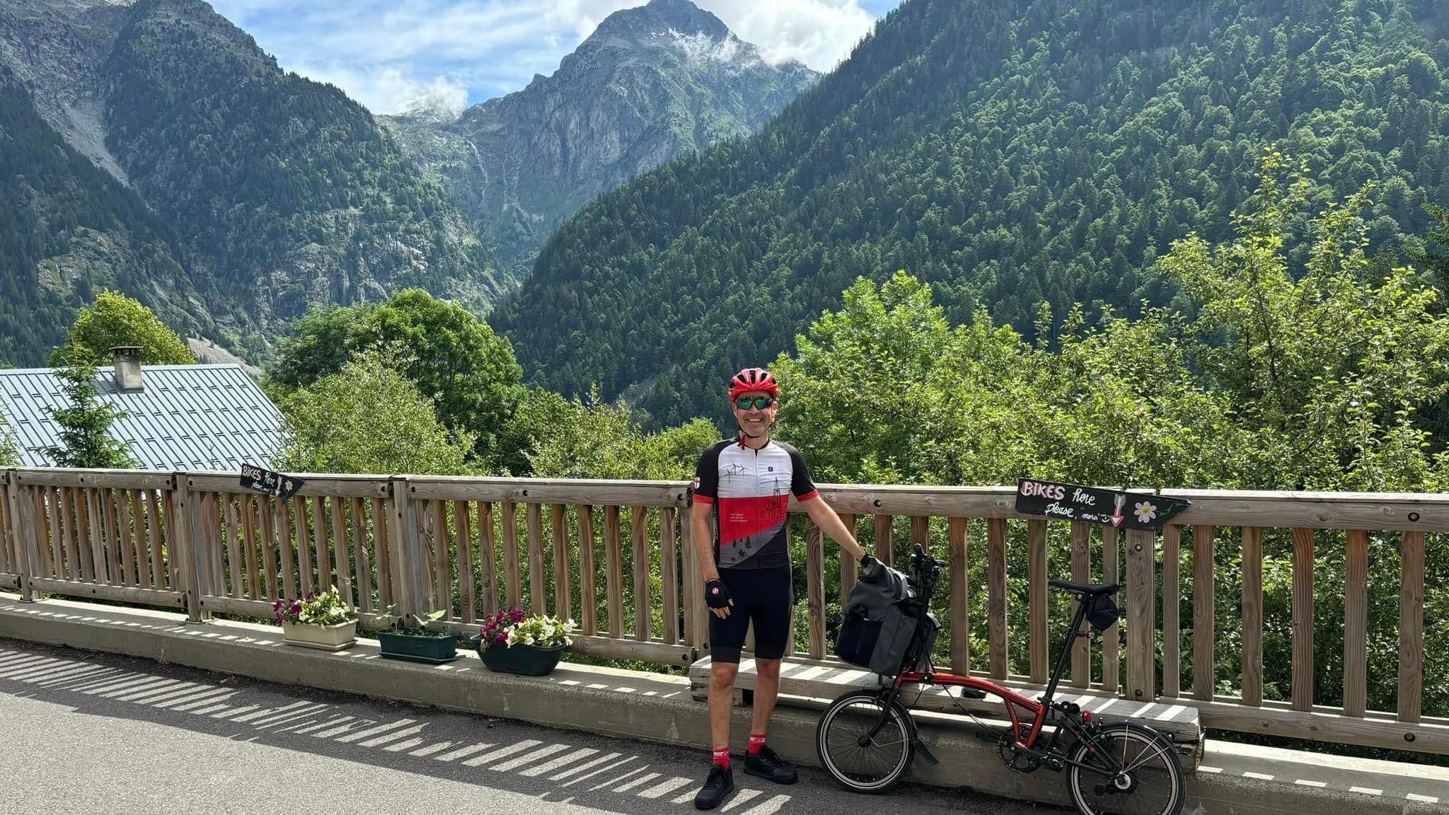 A smiling man in cycle gear with a portable city bike in front of a specatcular mountain range