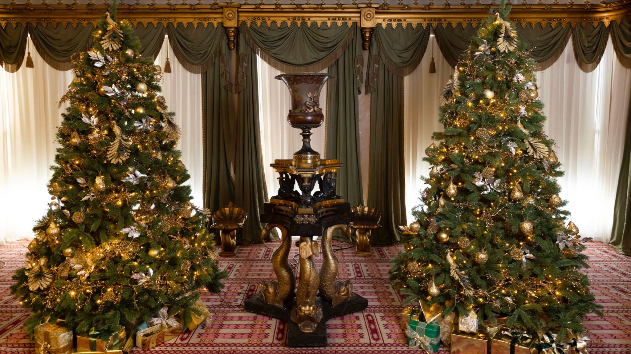 Two Christmas trees next to each other covered in golden lights and decorations, and a black and gold ornament in the middle of them