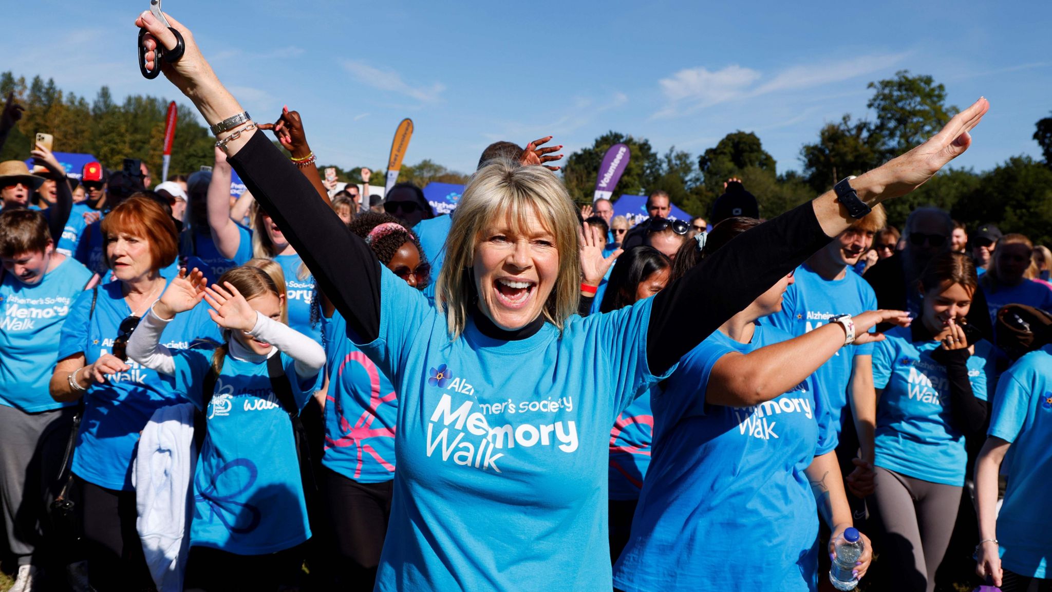 Ruth Langsford raising her hands and smiling on the memory walk surrounded by other walkers all wearing blue t-shirts with Memory Walk written on them