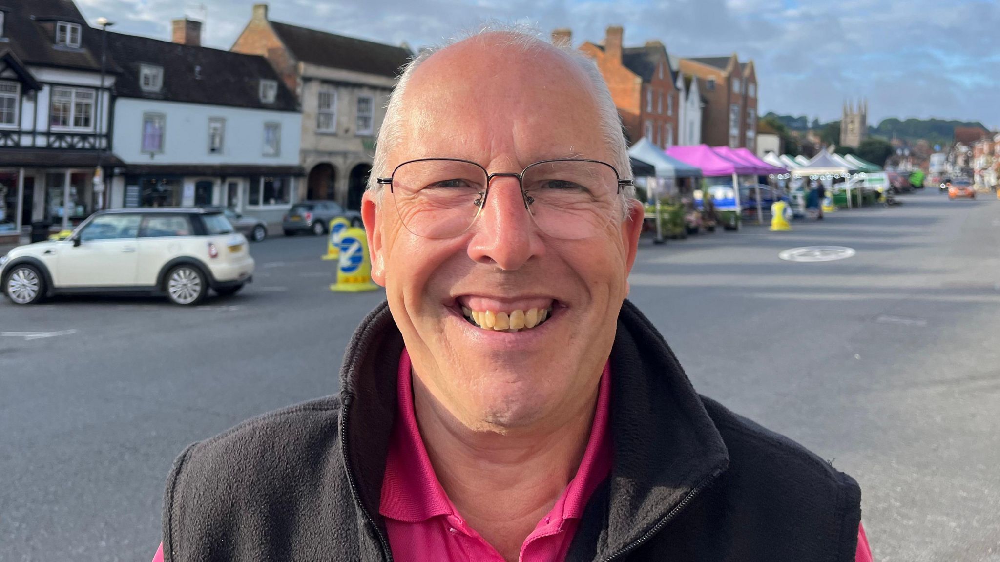 Head and shoulders photo of Bob Holman, who is wearing a pink t-shirt and black body warmer and glasses. He smiling and stood in front of market stalls in the background and a church can be seen in the distance.