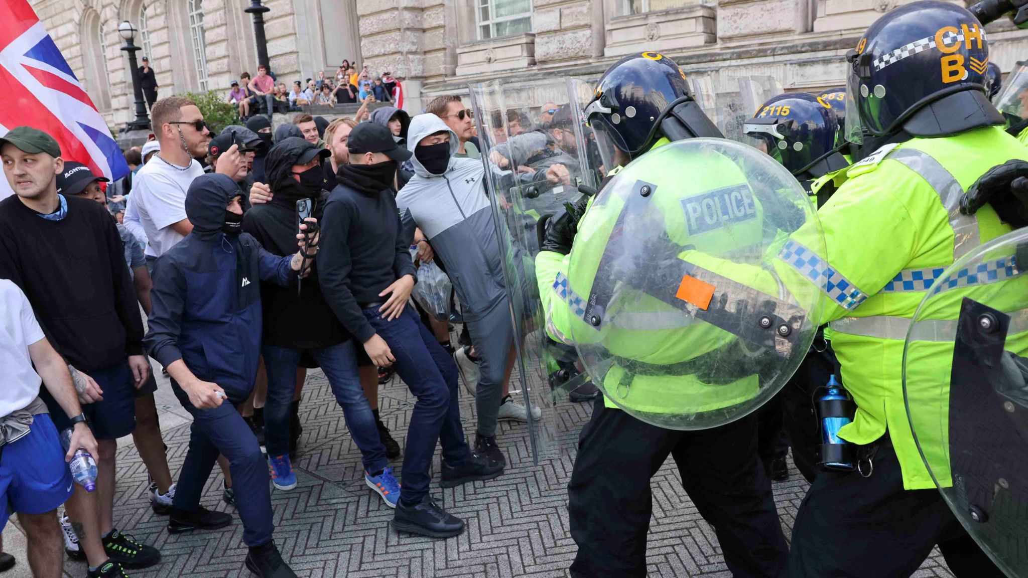 Protesters dressed in tracksuits, caps and face masks, clash with police in riot gear and holding shields outside Liverpool's Cunard building