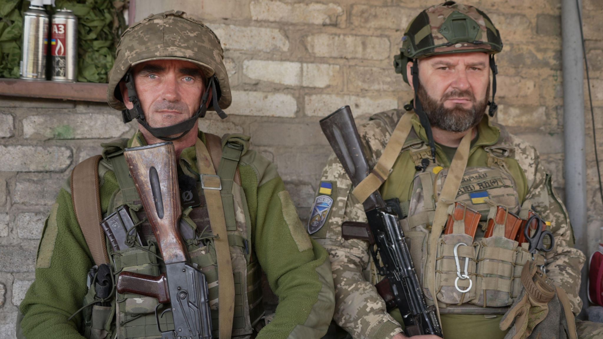 Two Ukrainian soldiers wearing helmets and carrying guns look into the camera lens