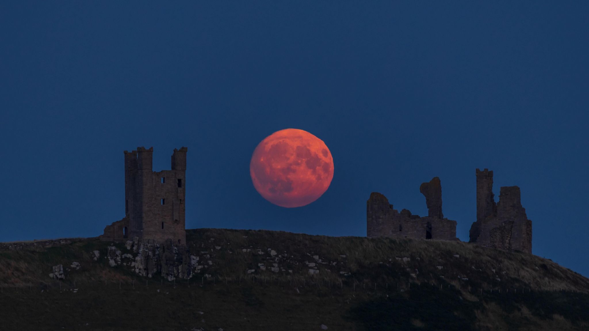 Going dark in the evening so we can just make out some castle ruins on top of a hill but behind in the background, the full moon rising low in the sky has a red tinge.