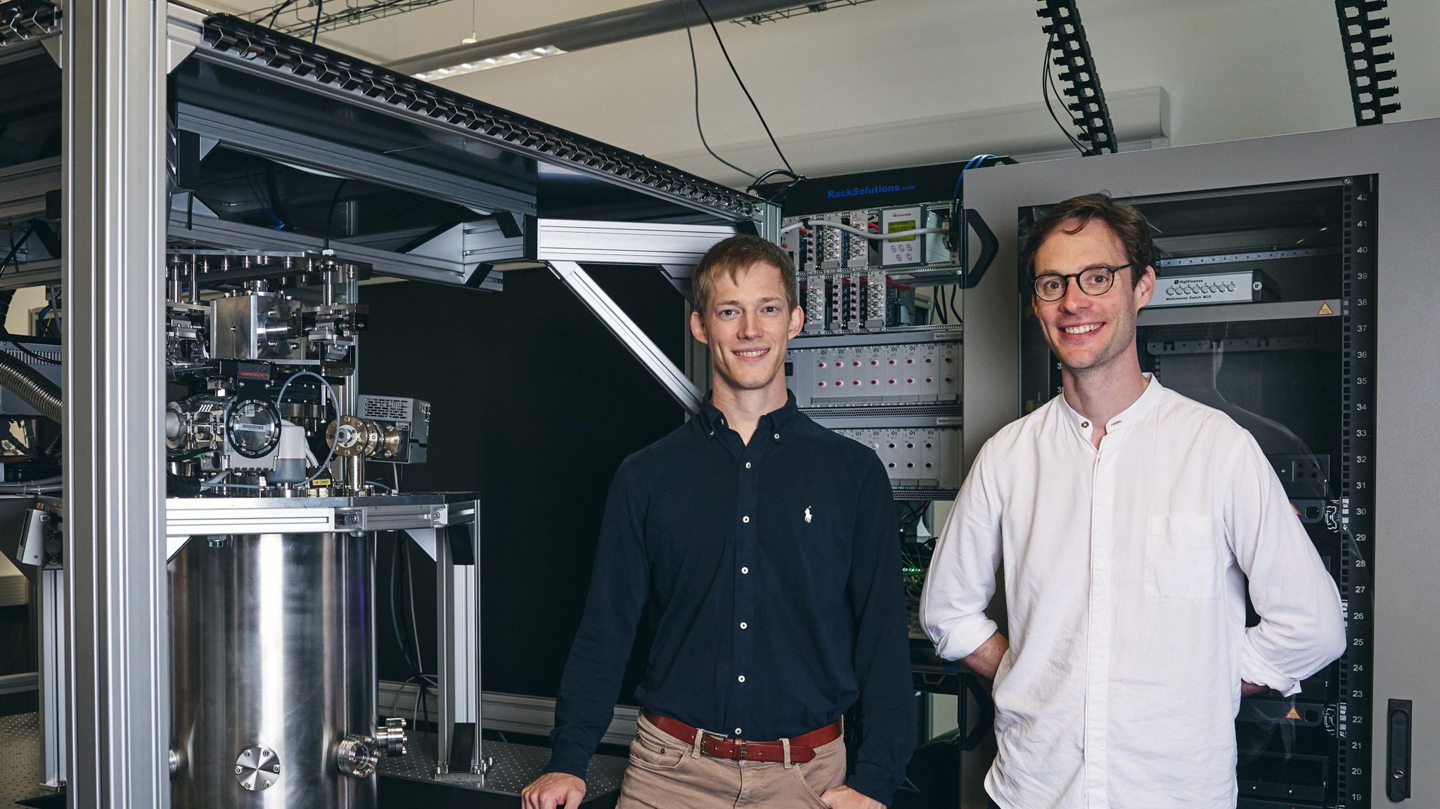 Two men stood in front of a science lab with a quantum chip in. One is fair-haired wearing a blue shirt, whilst the other has dark hair a white shirt and glasses