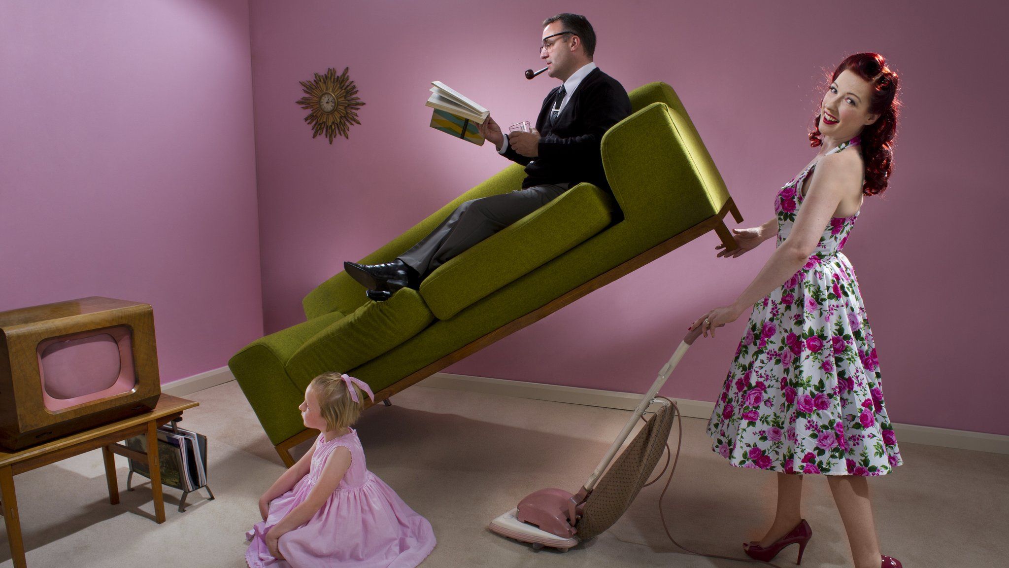 1950s stereotypical housewife uses vacuum cleaner while holding up her husband who's lying on sofa