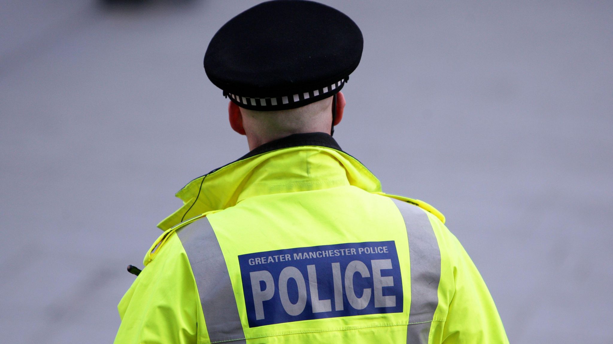 A police officer faces away from the camera, wearing a hi-vis jacket with 'Greater Manchester Police' written on it across a blue stripe