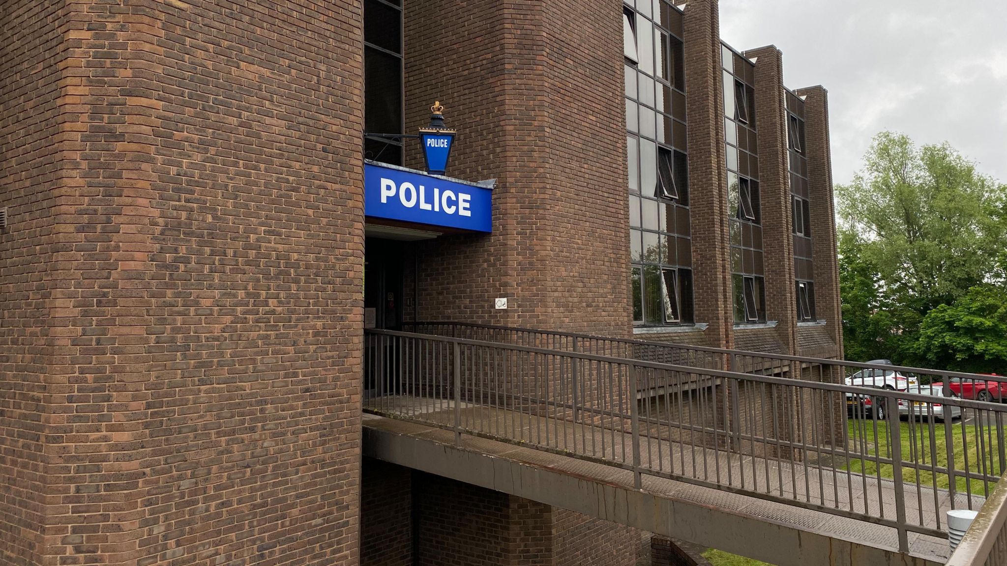 A brown brick building with a metal bridge leading to a door. Below the bridge is green grass. Above the door a large blue sign with white writing reads "police"