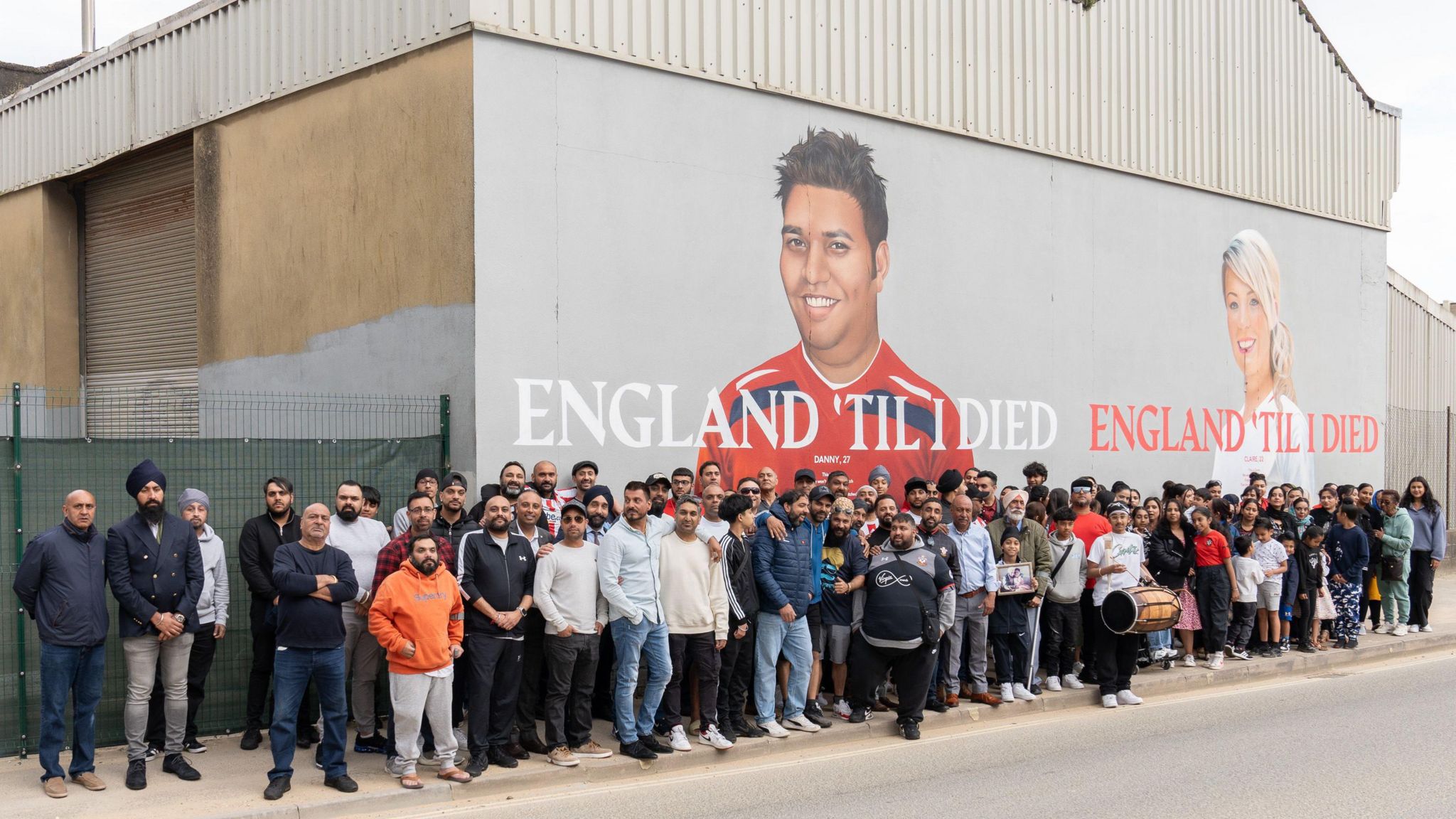 A large group of around 80 people standing in front of the mural, which depicts a young man and a young woman with the slogan 'England til I died'