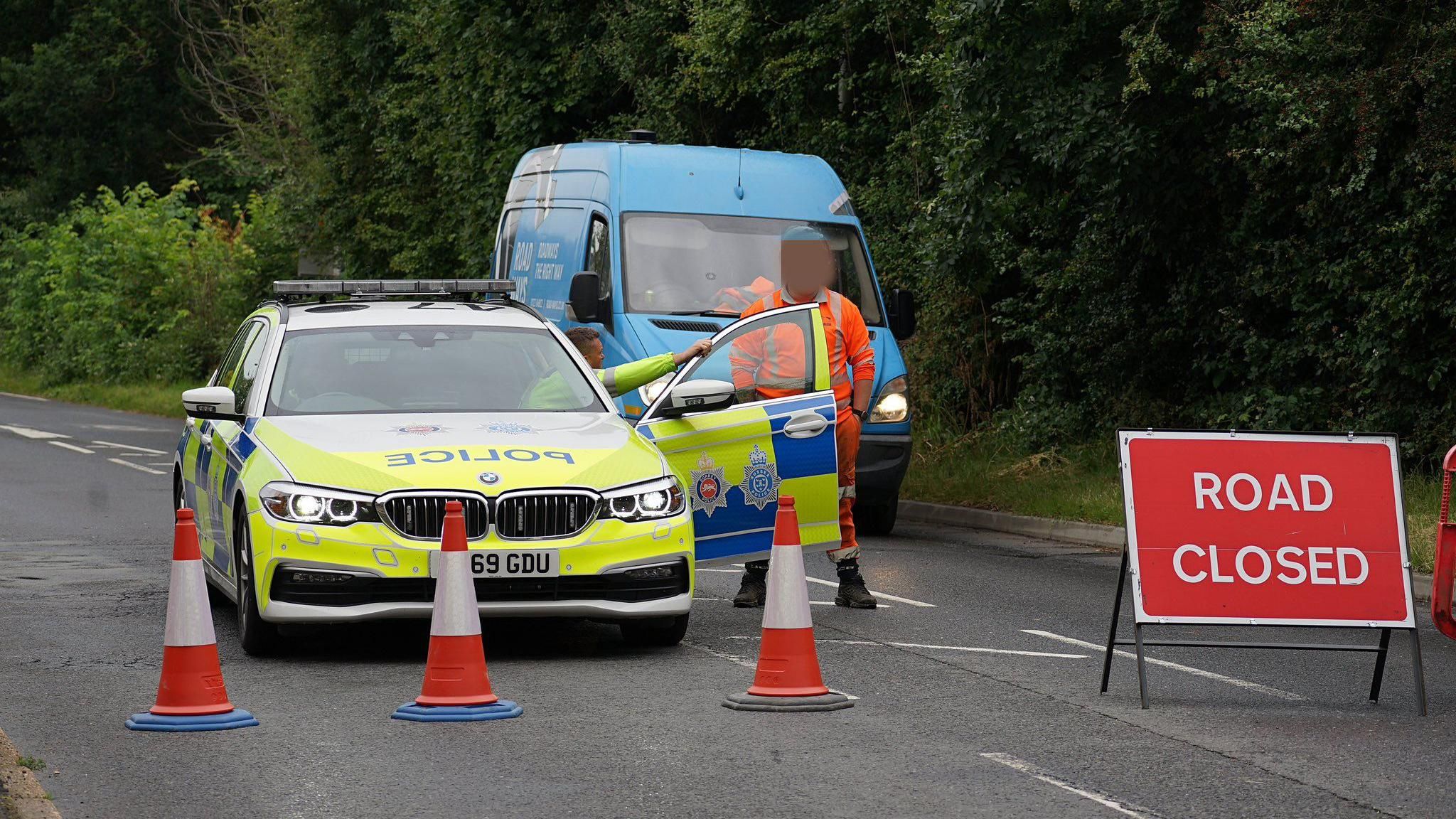 A police car behind three orange traffic cones next to a red sign which reads Road Closed. A man in an orange high-vis outfit is stood in front of a blue van