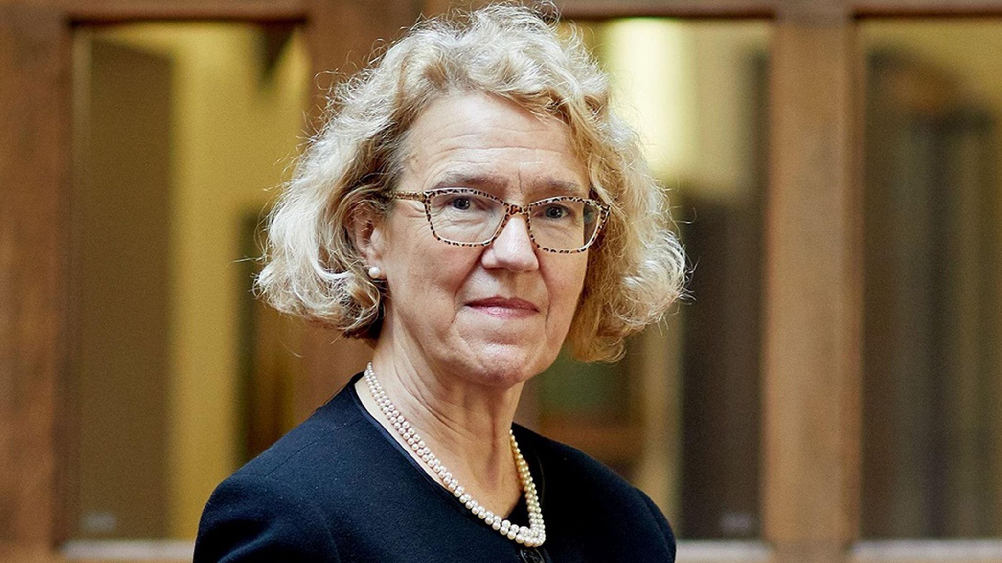 Lady Justice Thirlwall, who has curly blonde hair, smiles at the camera wearing turtle shell glasses, a dark blue blazer and a pearl necklace 