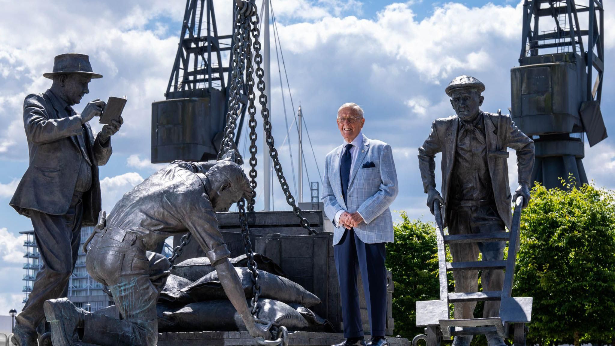 Johnny Ringwood, wearing a light blue suit jacket with navy blue trousers and tie, standing with the monument of three dockworkers at the Royal Docks in London