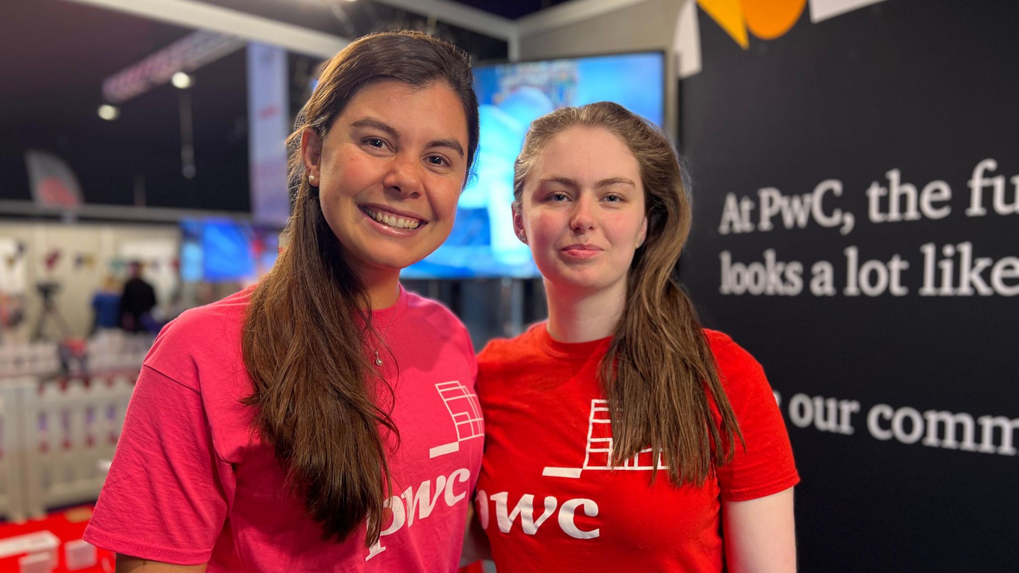 Ruth and Olivia smile at the camera wearing PwC tops and there is a PwC banner behind them