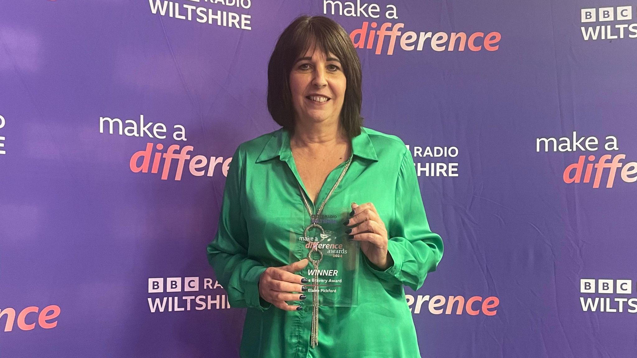 Elaine wearing a green blouse and holding her award in front of the BBC Radio Wiltshire purple banner