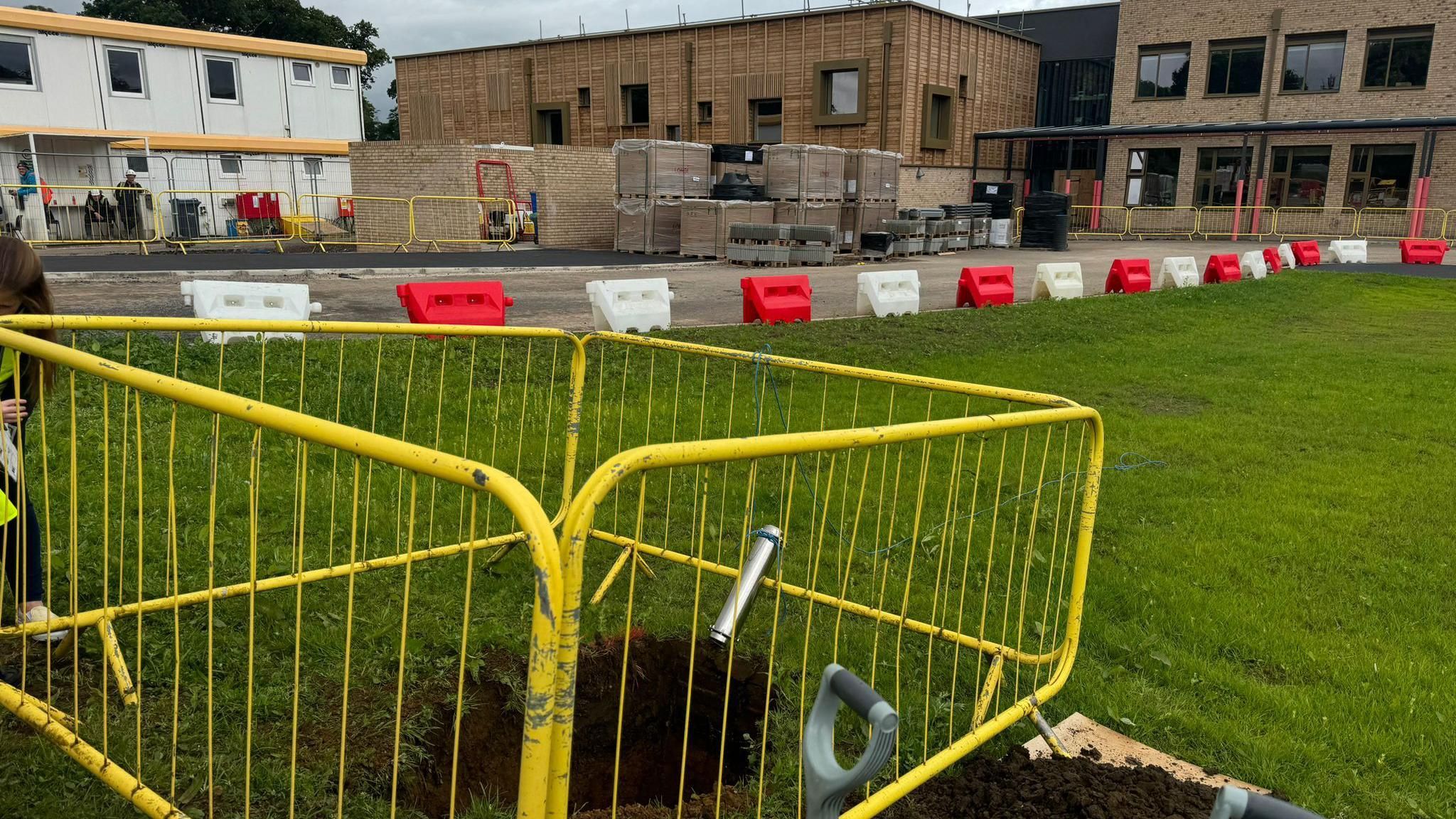 Silverwood School's Rowde campus in Wiltshire. Construction work can be seen next to existing buildings in the background. There is a bright yellow metal fence cordoning off a hole in the ground, where the time capsule is buried. 