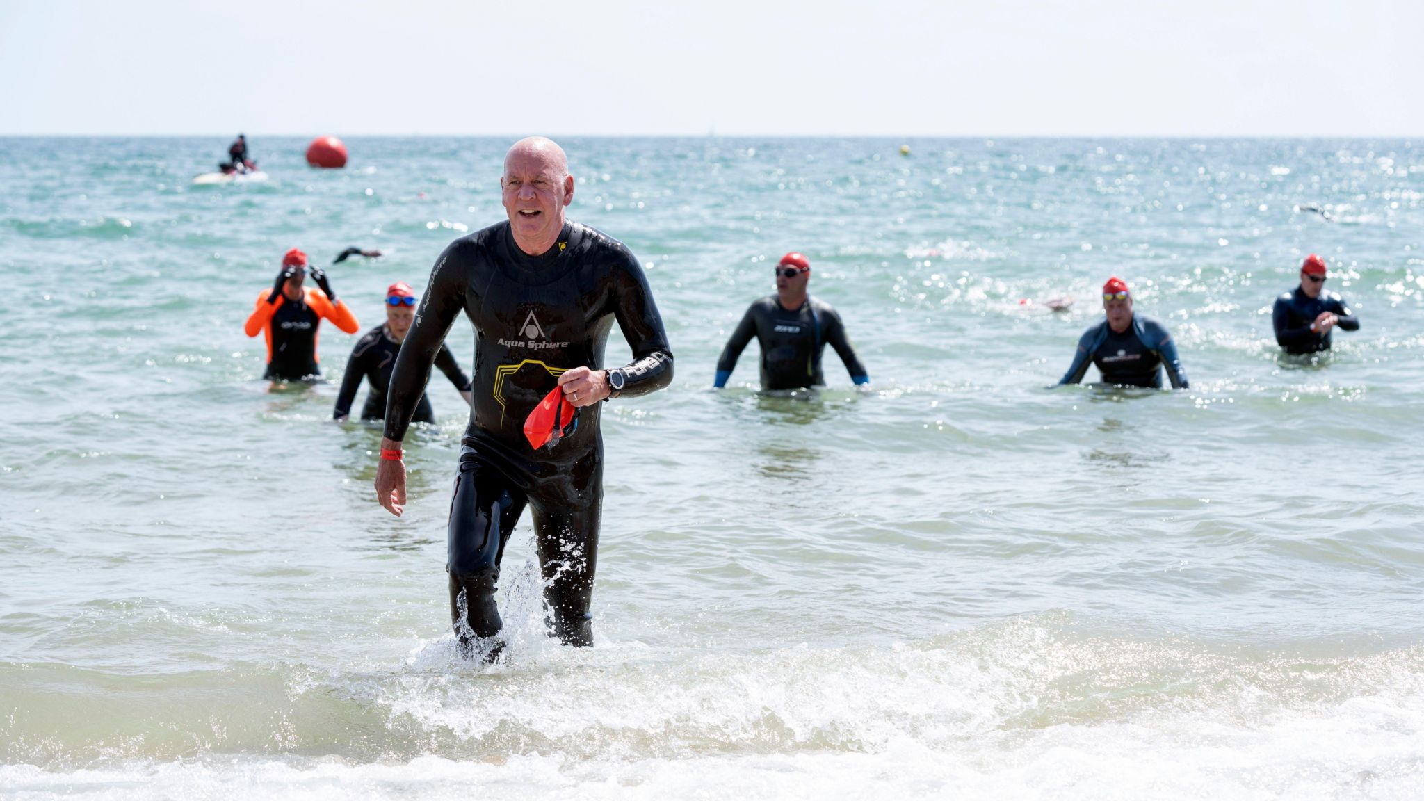 A group of swimmers wading out of the sea. One man can be seen at the front of the group holding his swim cap and goggles after removing them.