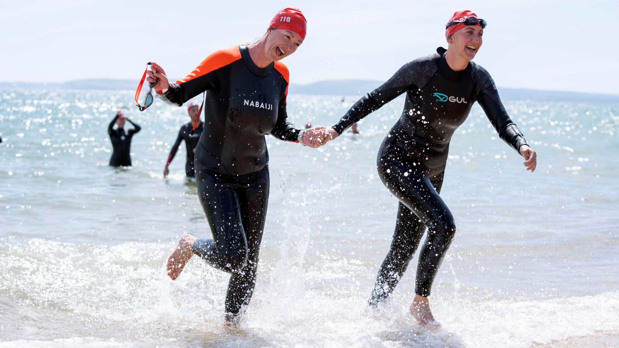 Two female swimmers running from the sea, splashing water, smiling and laughing in their wet suits and swim caps after removing their goggles.