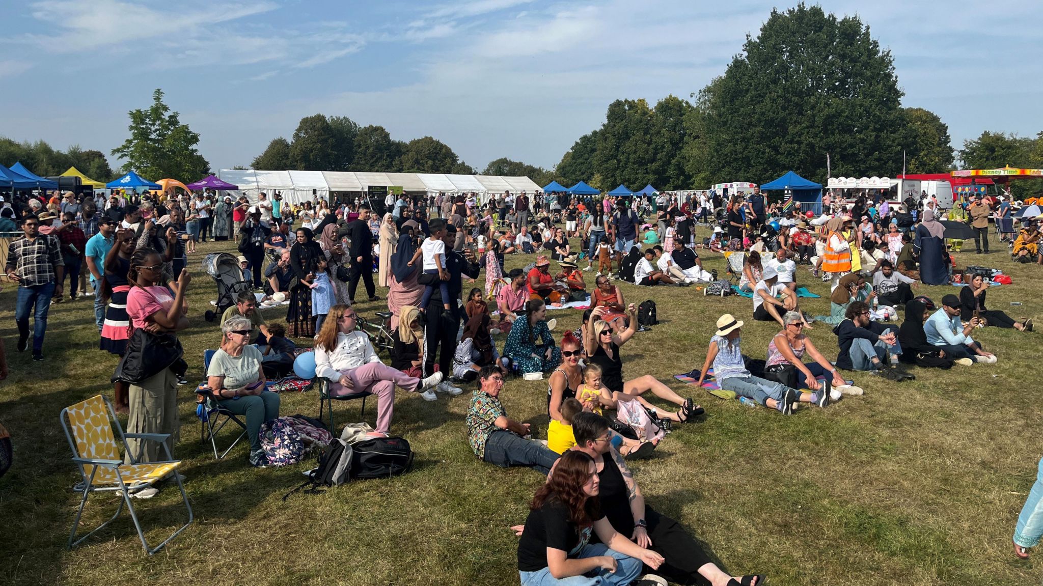 Crowds of people of different ethnicities sitting and standing in a park, looking towards a stage