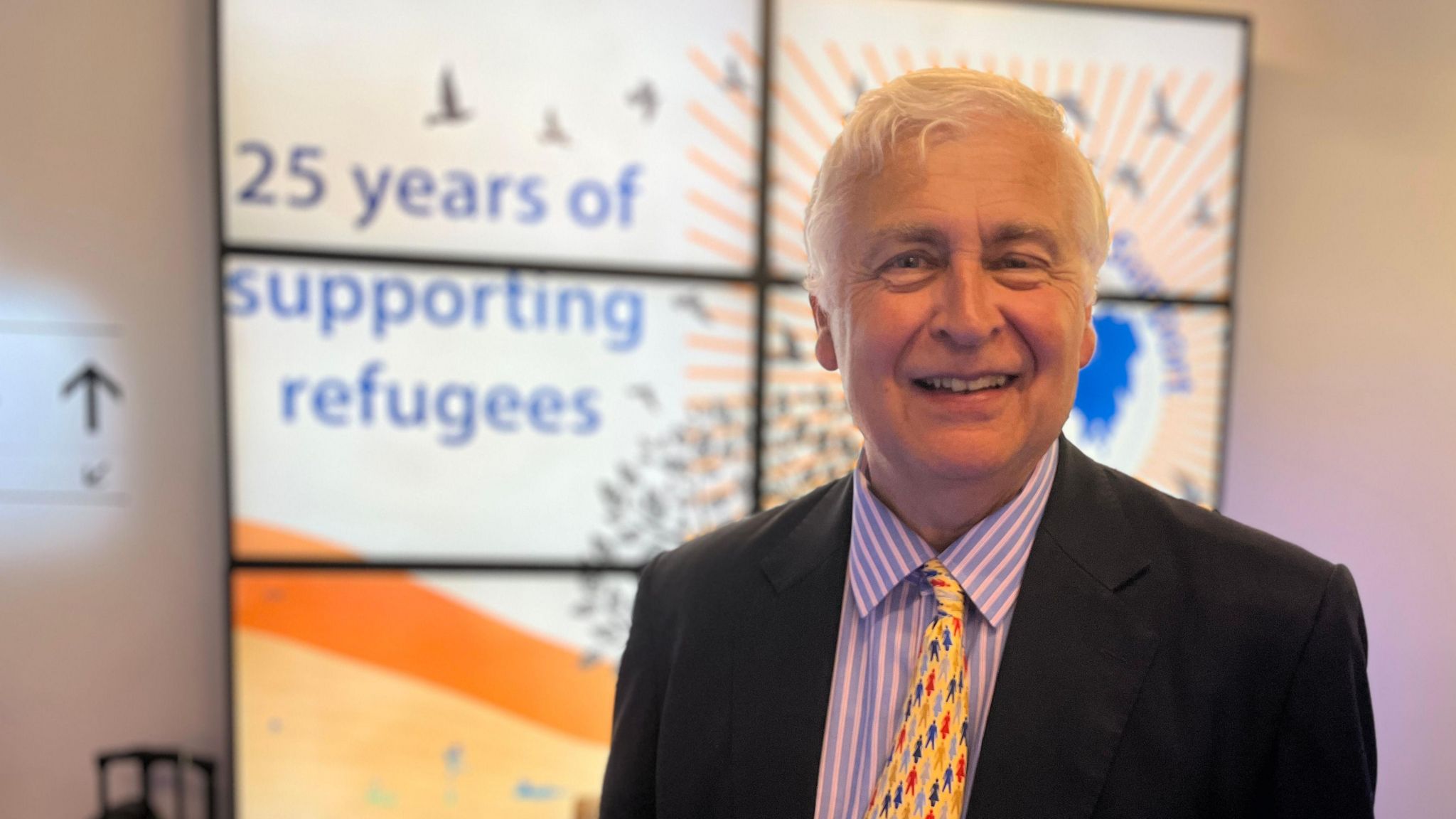 Sir Nick Young wearing a suit and tie stands in front of a banner saying 25 years of supporting refugees