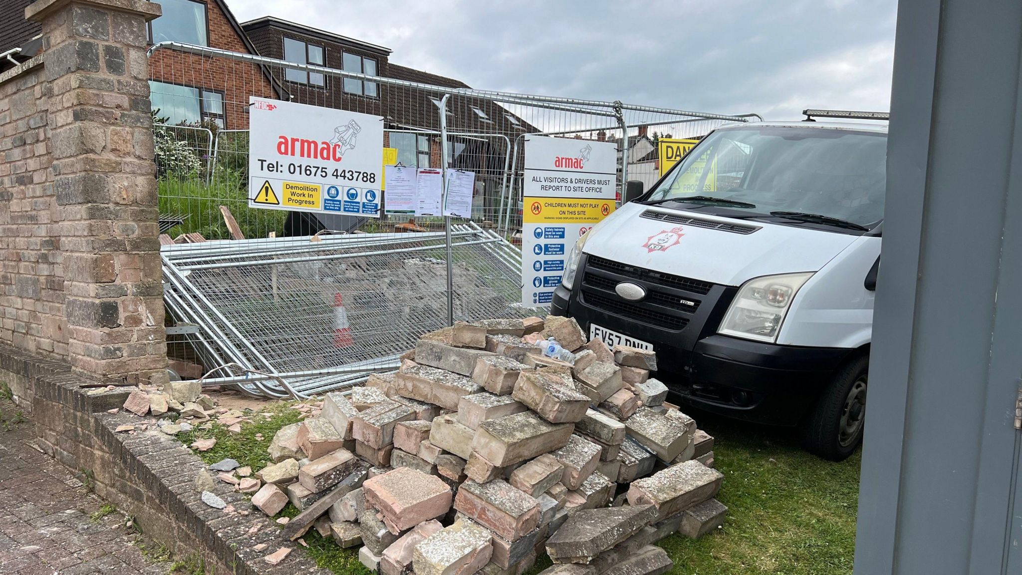 Picture shows a white van outside a house, which is behind large silver fencing. A pile of bricks from the demolished building is visible in the foreground. 