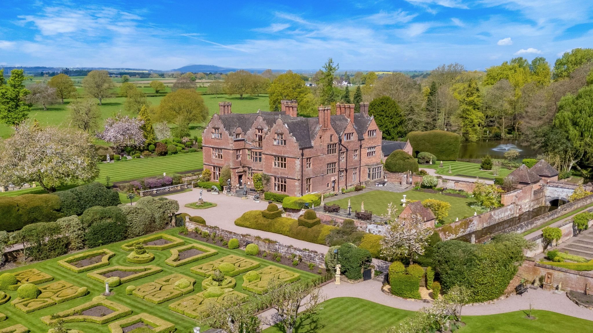 A large, brown brick manor house sits surrounded by green gardens and hedges. In the background are green fields and trees, the sky is blue.