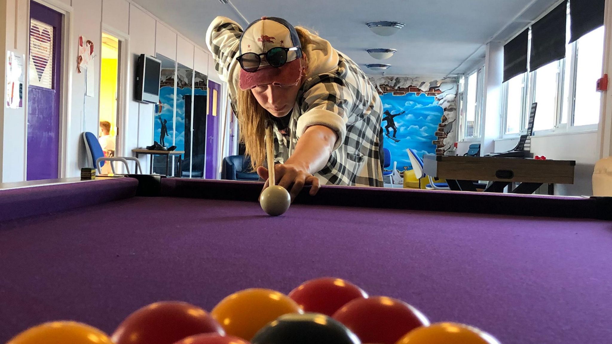 A young man with long hair and a cap lines up a break on a pool table