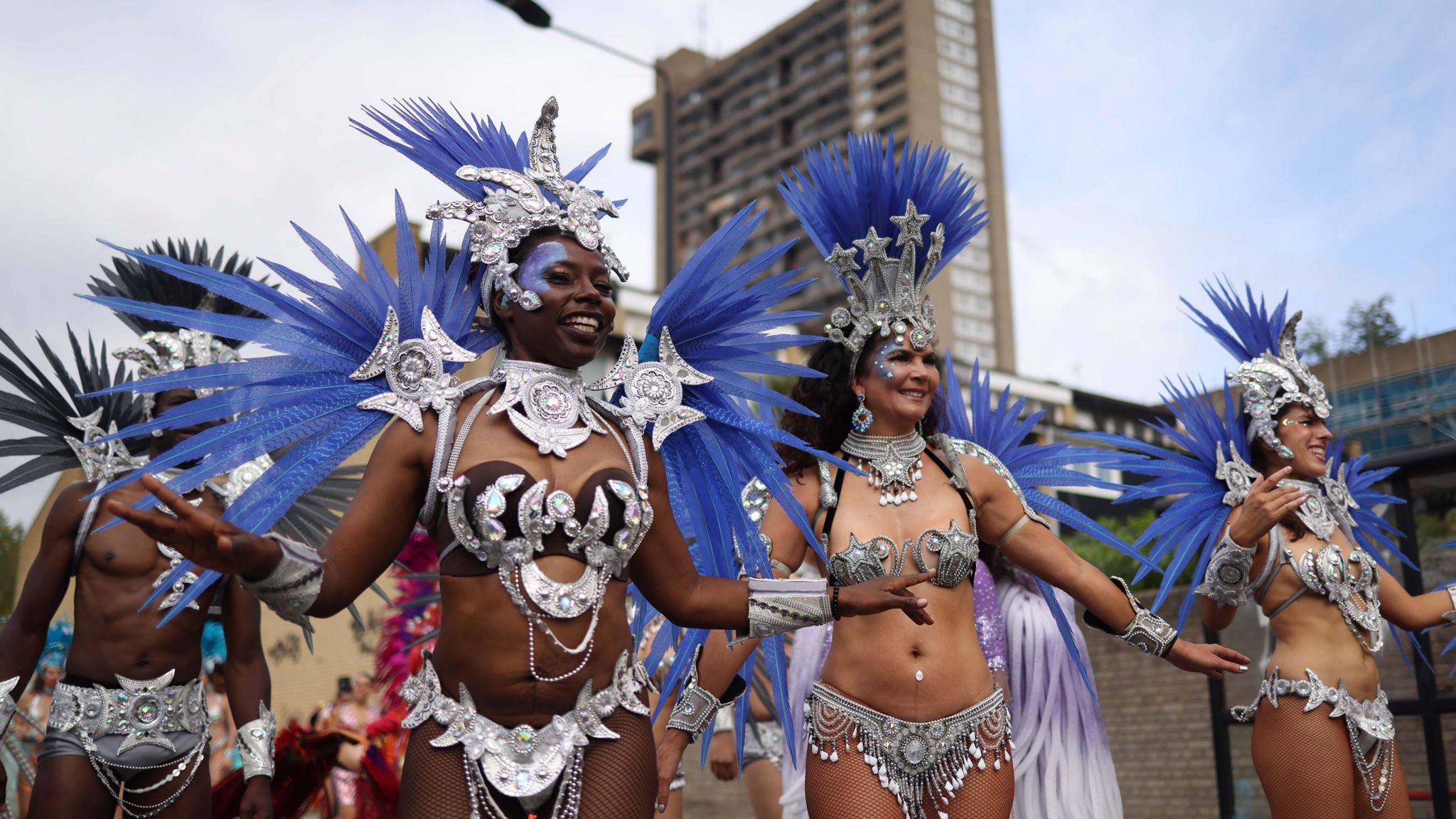 Dancers at Notting Hill carnival wearing silver, metallic and jewel encrusted bikinis, trunks and their headpieces are silver with long blue feathers/leaves.