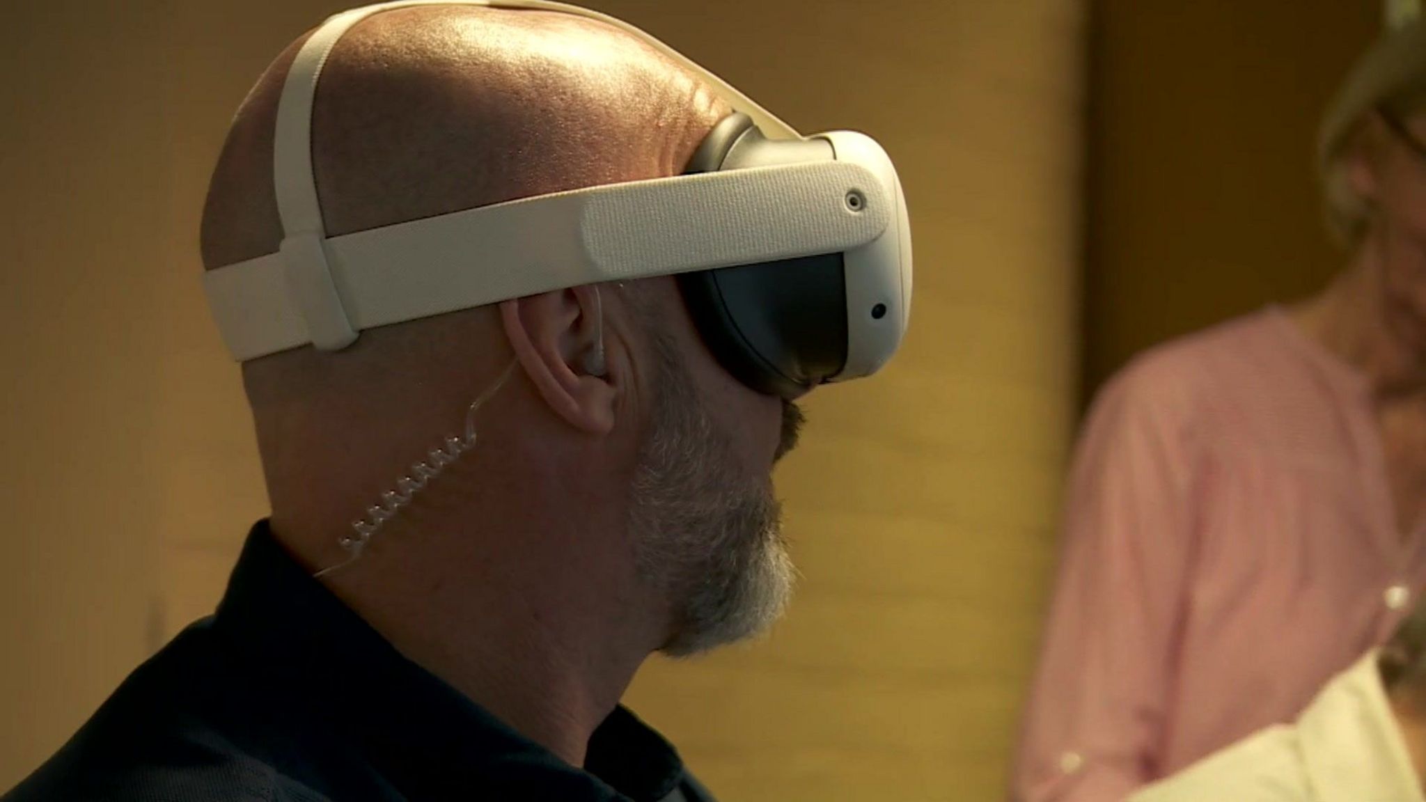 A man wearing a white virtual reality set. He is in the room with other people, only his left profile is visible as he is turned away from the camera