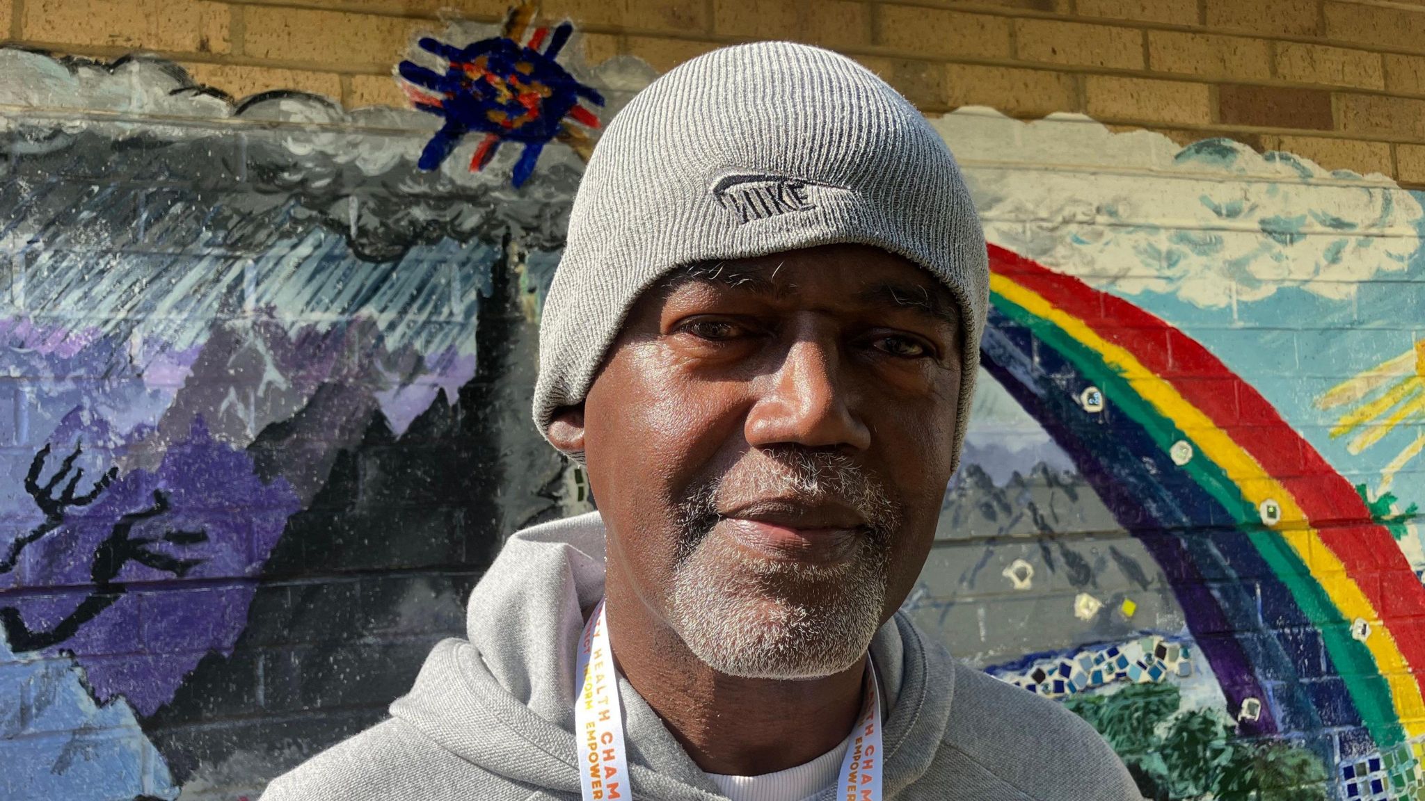 Errol Masters is wearing a grey Nike beanie hat, is wearing a grey hooded top and a white and orange lanyard. He's standing in front of a painted mural, which includes a rainbow on the right of the picture, and a stormy scene on the left 