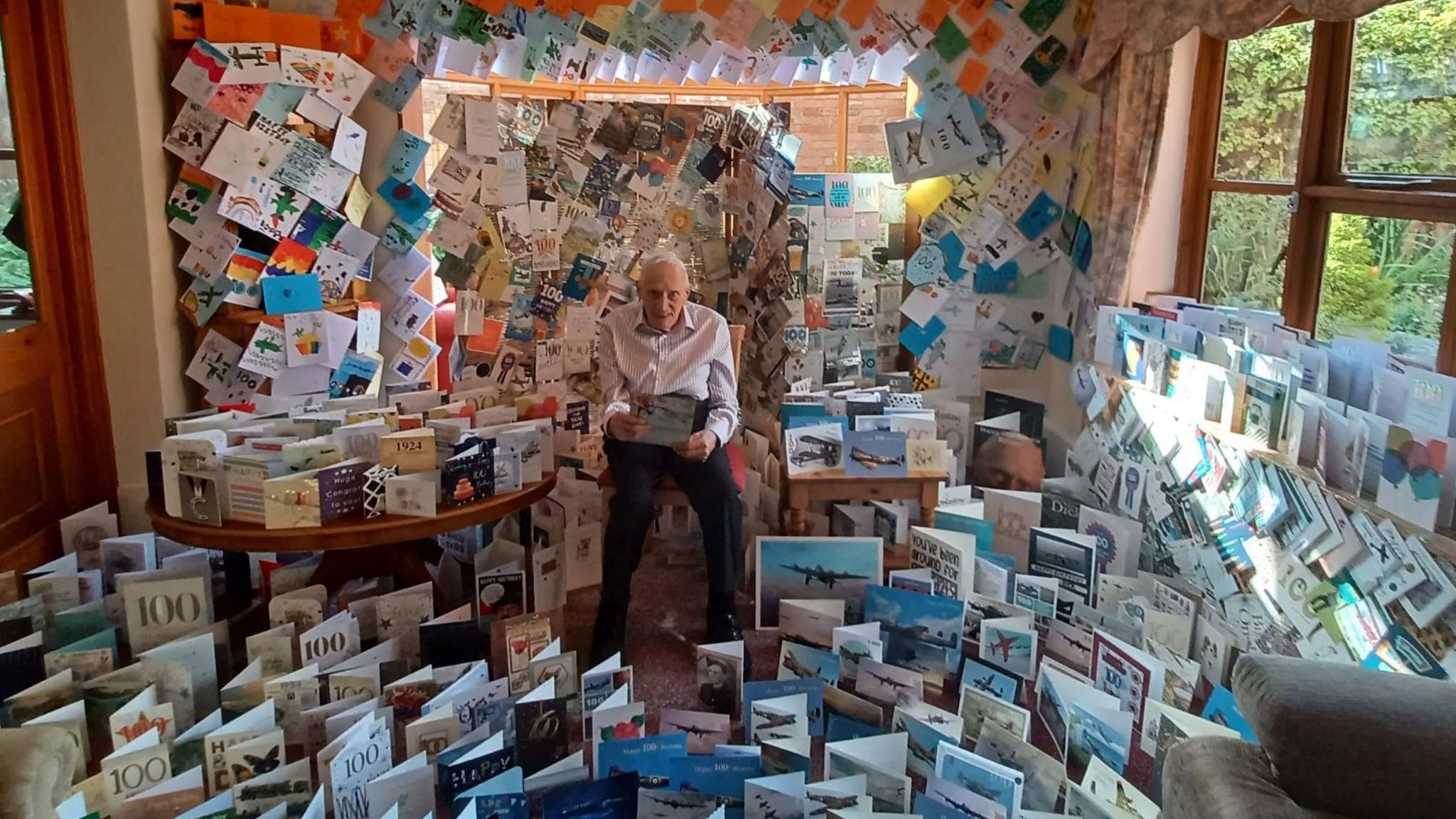 An old man in a white shirt and black trousers holds a card in a room that is filled with birthday cards - on the floor and covering every inch of the walls