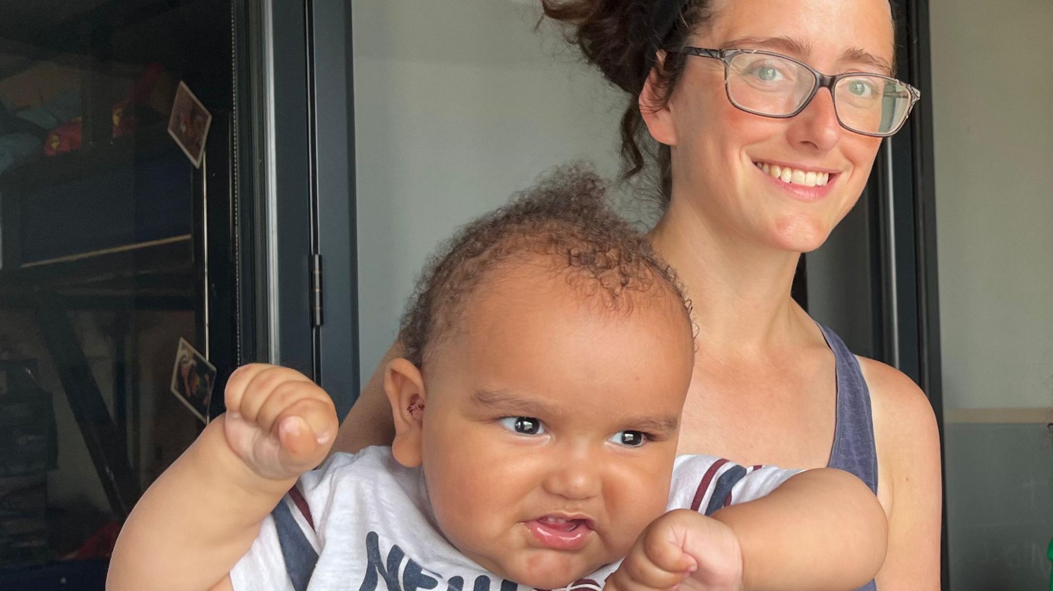 Thea, a young woman with brown hair and glasses, and with her baby son waving his arms in the foreground of the photo