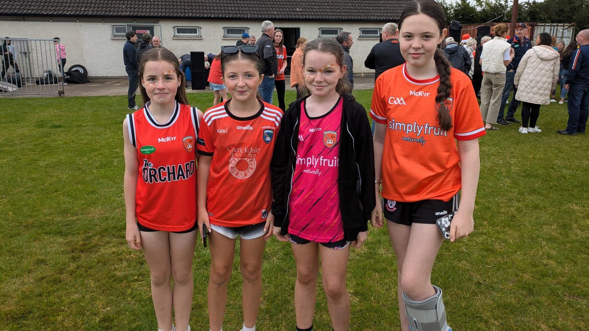 Four girls wearing Armagh jersey's standing on a GAA pitch, one is wearing and ankle boot