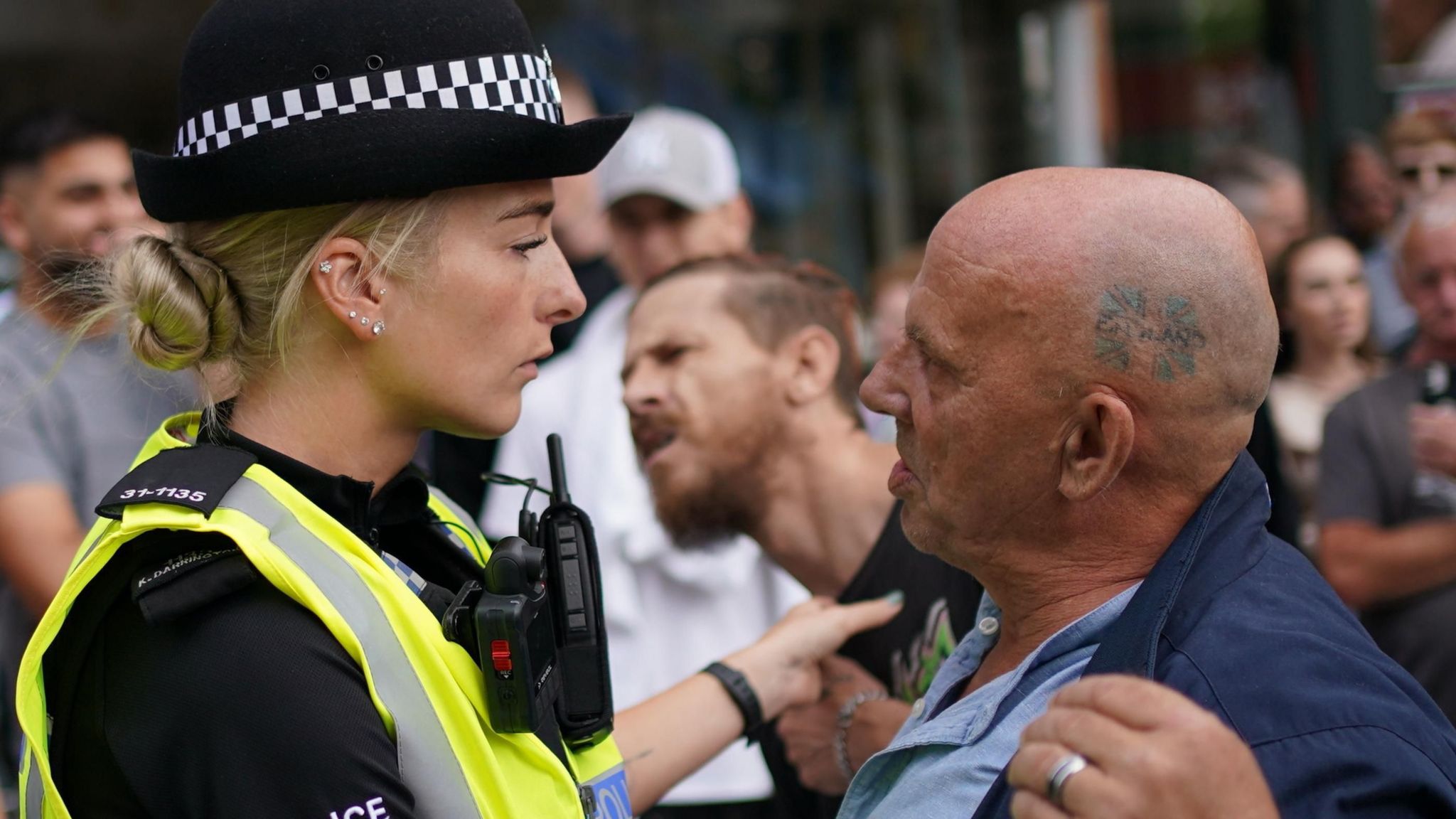 A woman police officer stands face to face with a shorter bald man with an England tattoo on the side of his head.