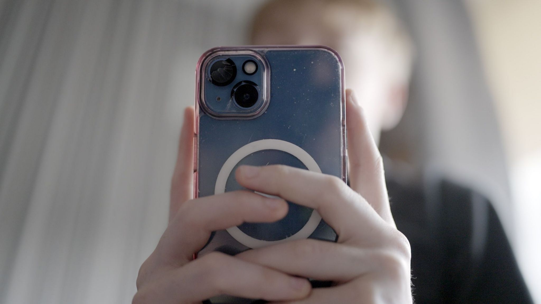 A close-up image of a teenage boy holding an iPhone with both hands. The phone and its camera lenses dominate the image, while the boys face is hidden and out of focus