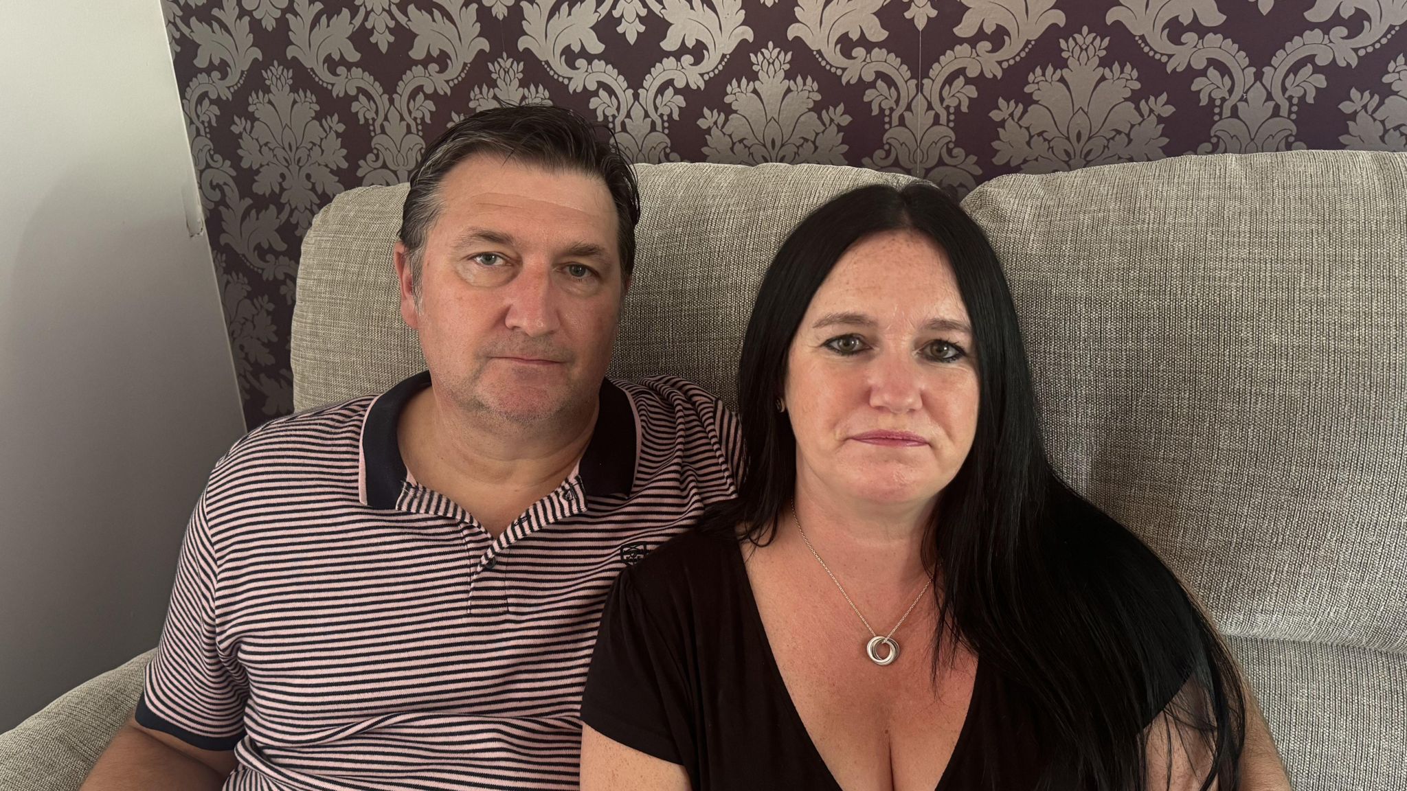 Nick and Vicky look directly at the camera, photographed sitting on a beige sofa. Nick is wearing a red and white striped polo neck, and Vicky is in a black T-shirt and wearing a silver necklace. 