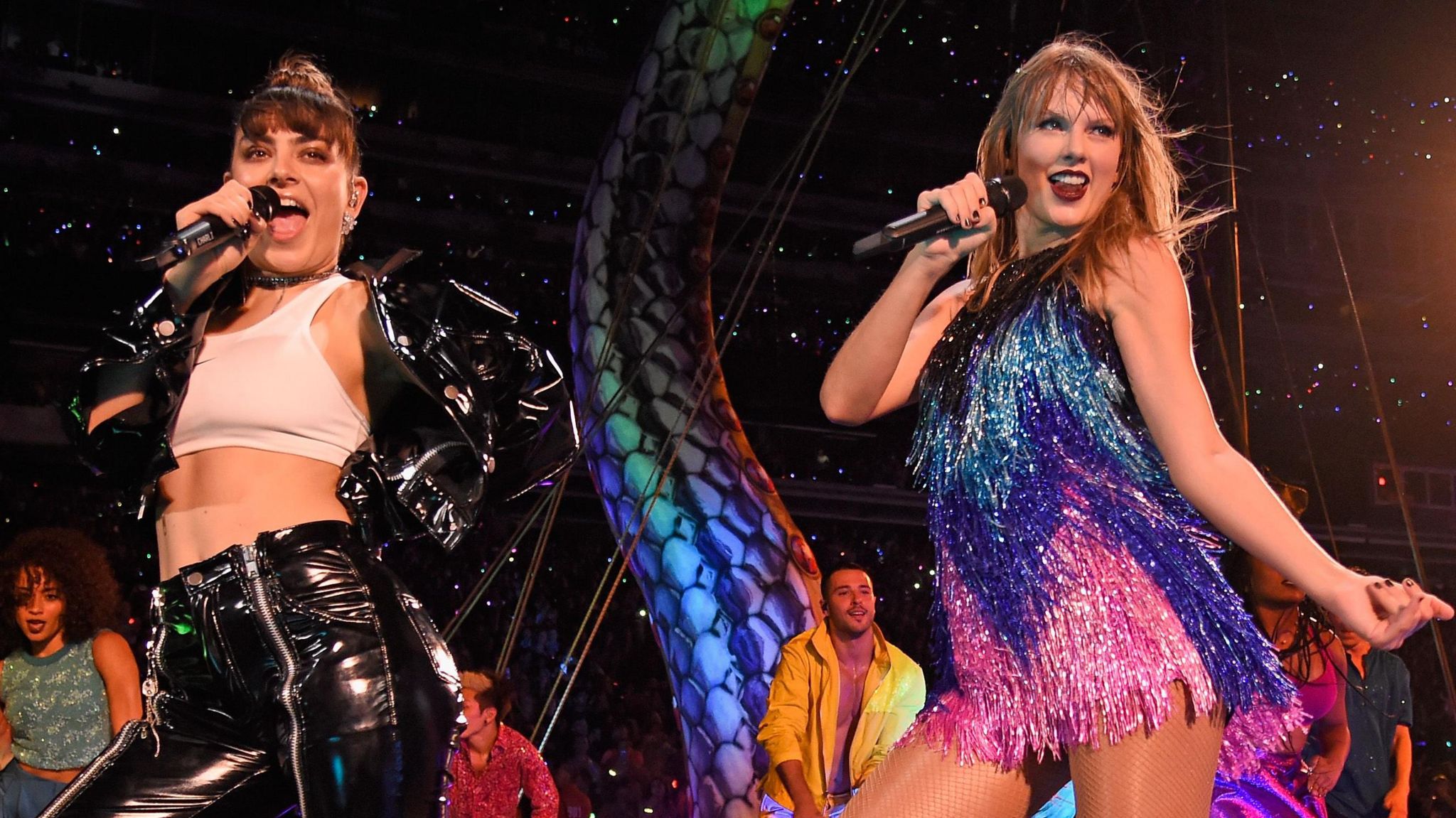 Charli XCX and Taylor Swift on stage together as part of Taylor's Reputation tour in 2018. Charli wears her dark hair in a top knot and wears a white crop top paired with a patent black jacket and matching trousers. Taylor wears a sparkly mini dress in pink, blue, purple and black. She has long blonde hair and red lipstick. 