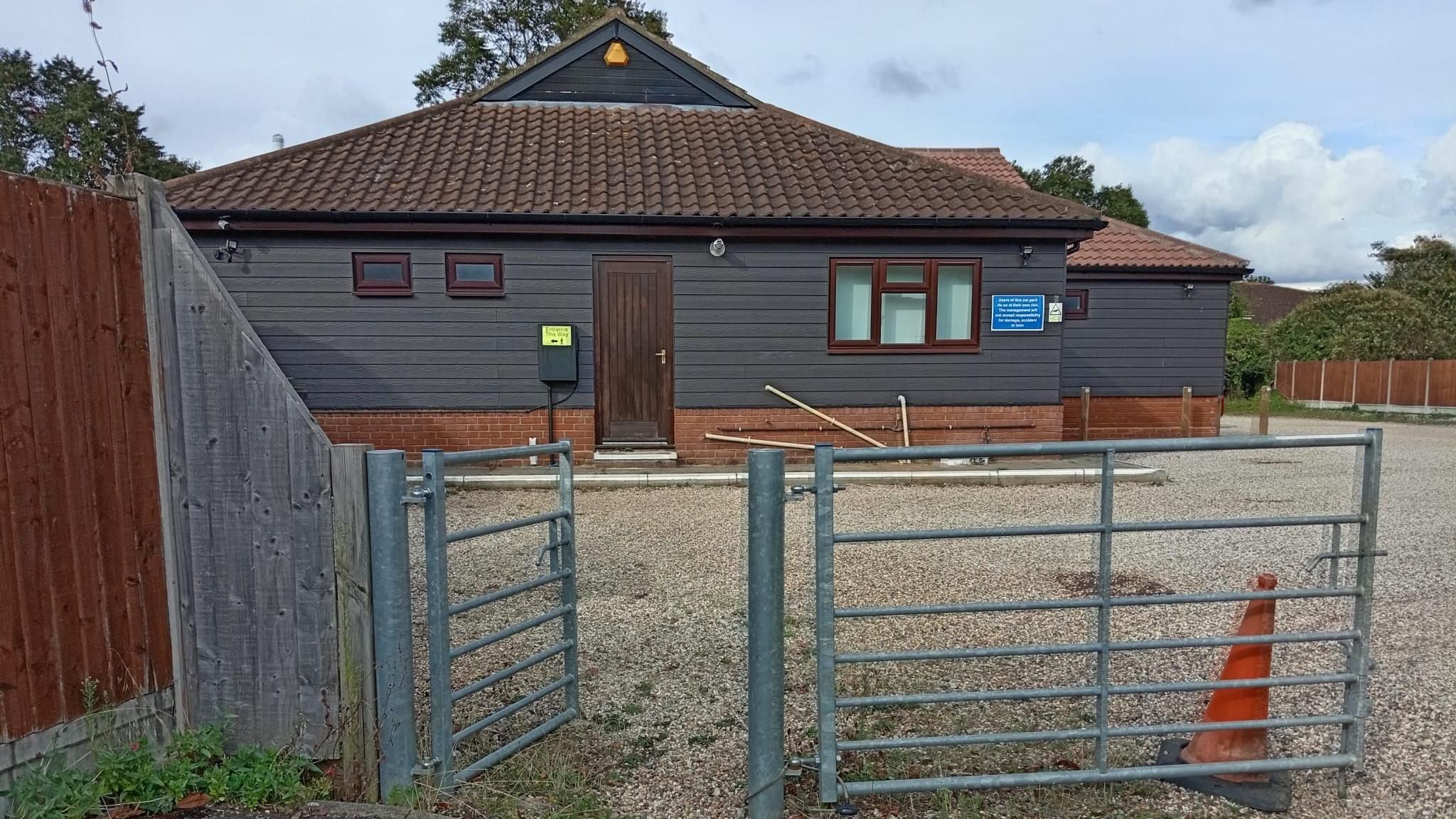 The entrance to RHP Bowls Club. It is a one-storey, black building with a brown roof. In the foreground is a metal gate.