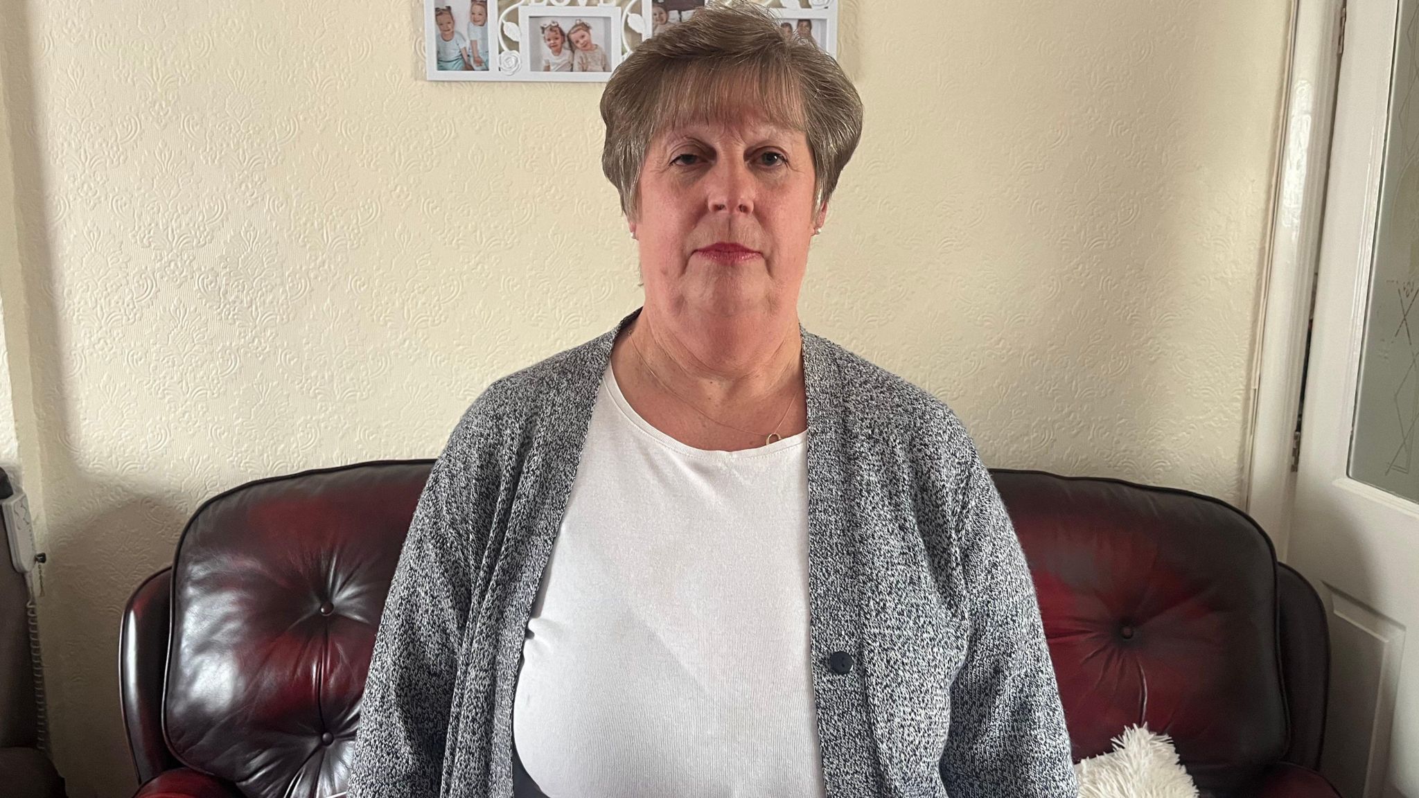 Jean Cross in her living room, with a dark brown sofa behind her and photos of family on the wall