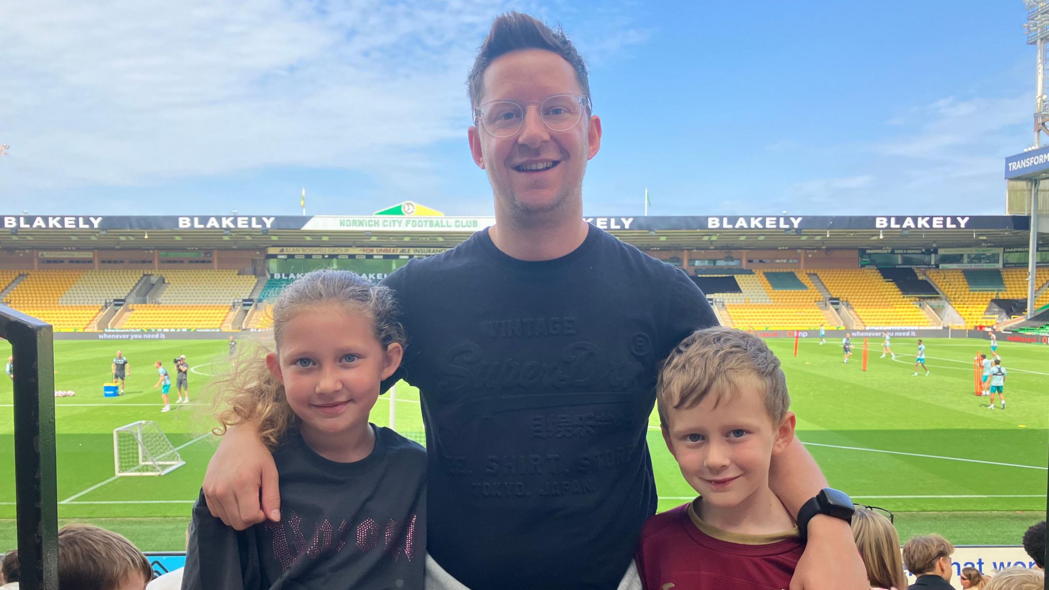 Simon with his children smiling at the camera, with the pitch behind