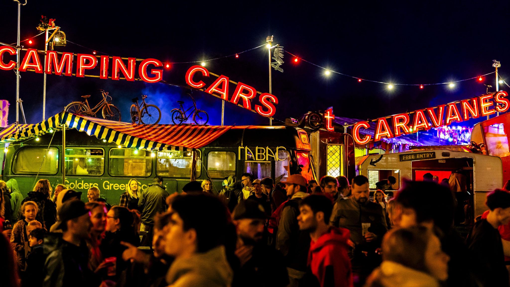 Dozens of people have gathered beside a repurposed bus and a caravan. Above them, neon red lights read "camping" "cars" and "caravanes". On top of the vehicles are bicycles. The night sky in the background is dark.