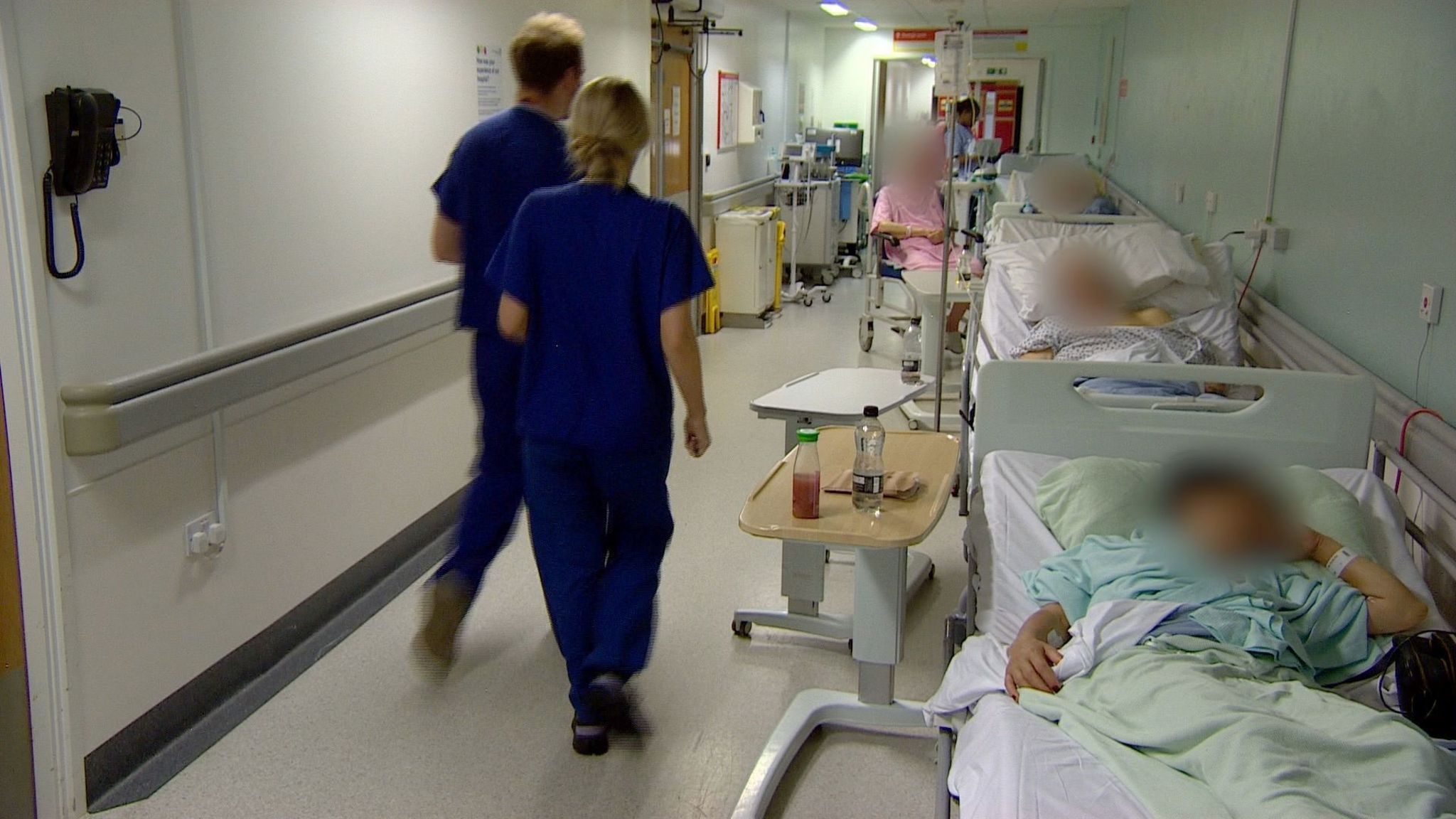 Patients lying in the corridor in beds on the righthand side of the photo, and on the left there are two nursing staff walking along. The patients' faces are all blurred out.